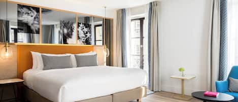 A modern hotel room with a large bed, a small table, a chair, and a mirror with artwork.