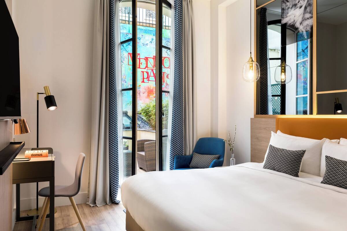 A hotel room with a bed, a desk, a chair, a lamp, and a view of the outside.