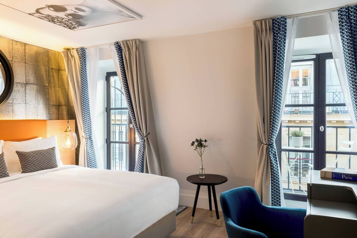 A hotel room with a large bed, a small round table with a vase of flowers, a blue chair, and a view of the city through a large window.