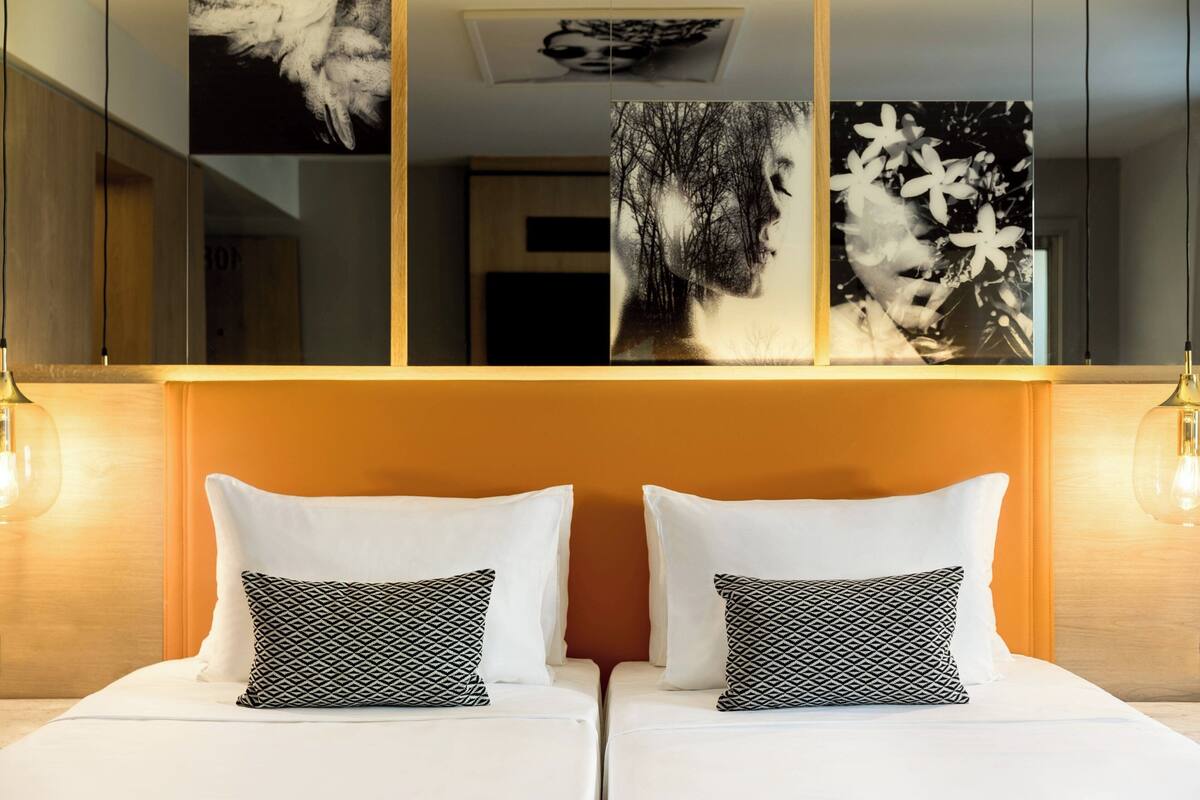 A hotel room with two beds, each with white bedding and patterned pillows. There are wall-mounted artworks above the beds.