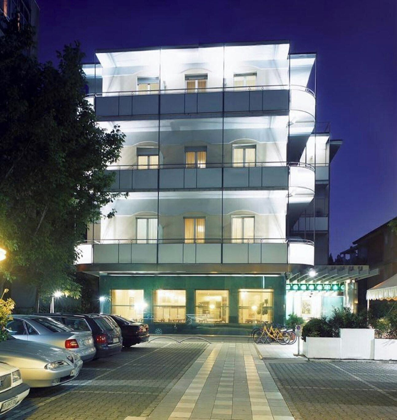 A multi-story hotel with a glass facade, a parking area with cars, and a well-lit entrance.