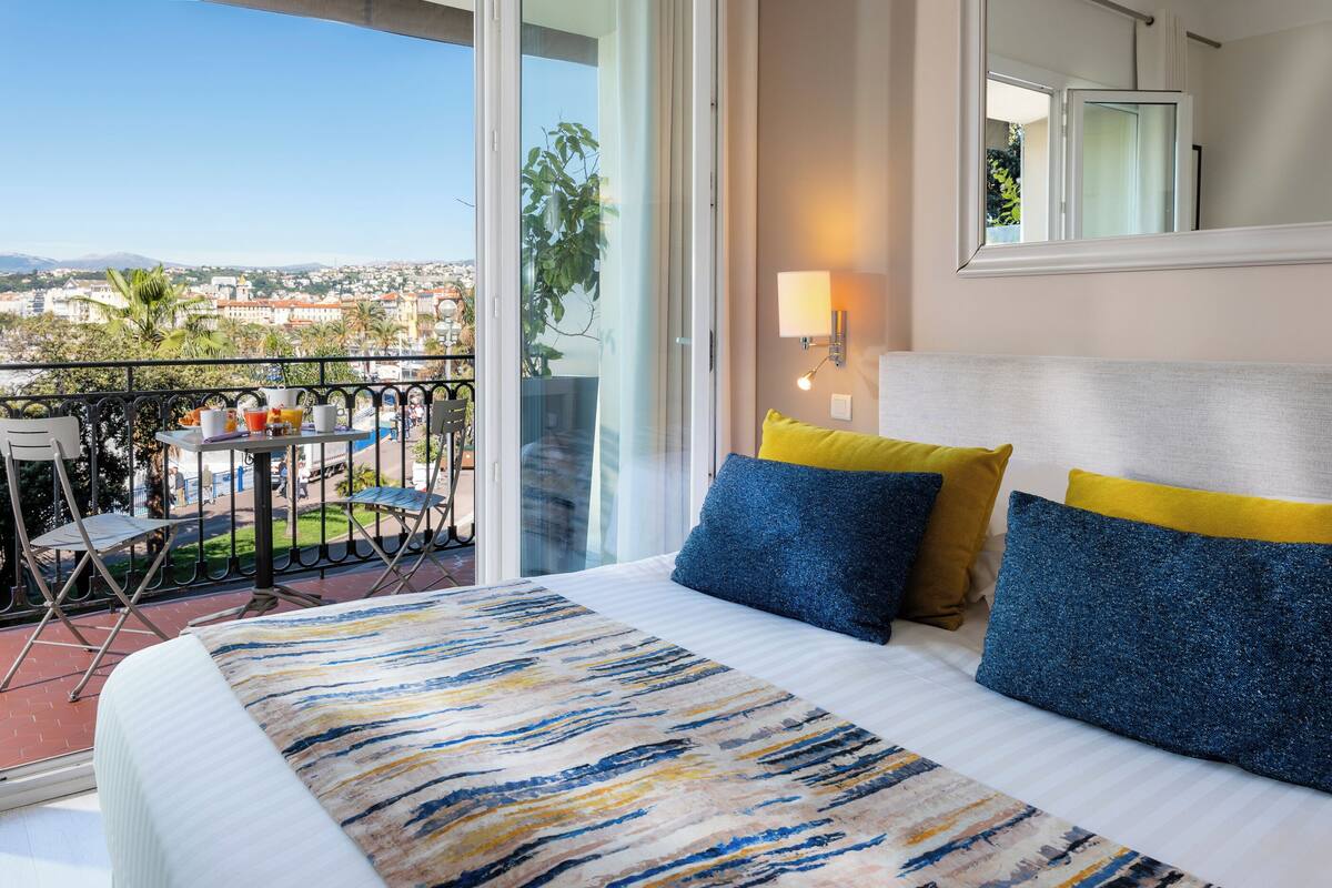 A hotel room with a balcony, a bed with blue and yellow pillows, and a city view.