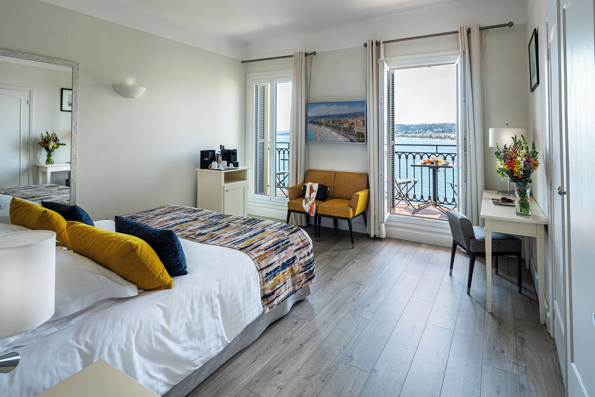A hotel room with a bed, a desk, a chair, a balcony with a view, and a painting on the wall.