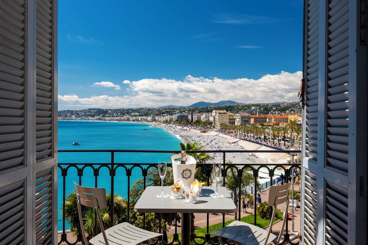 A balcony with a table set for two, overlooking a beach with a clear blue sea and a cityscape in the background.