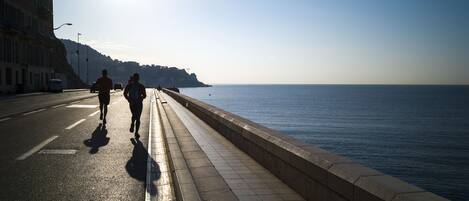 A coastal walkway with a paved path alongside a body of water, buildings on one side, and a clear sky.