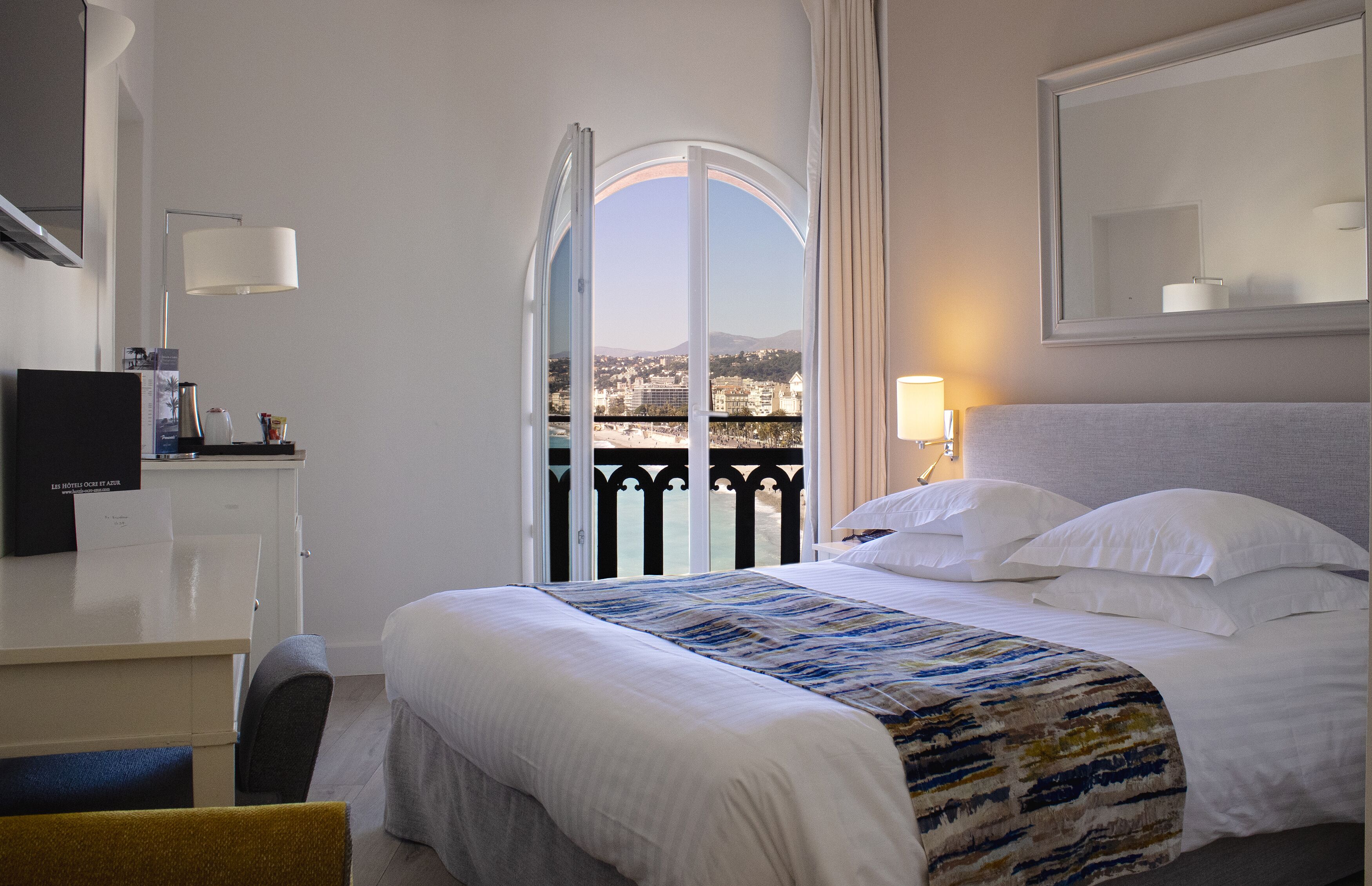 A hotel room with a large bed, a desk with a computer, a chair, a TV, and a view of the city through a balcony door.