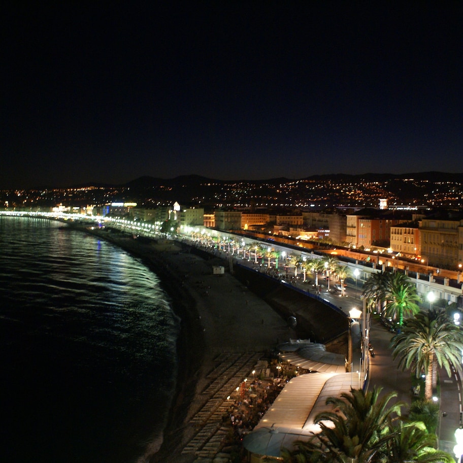 A coastal cityscape at night with a beachfront promenade, illuminated buildings, and a calm sea.
