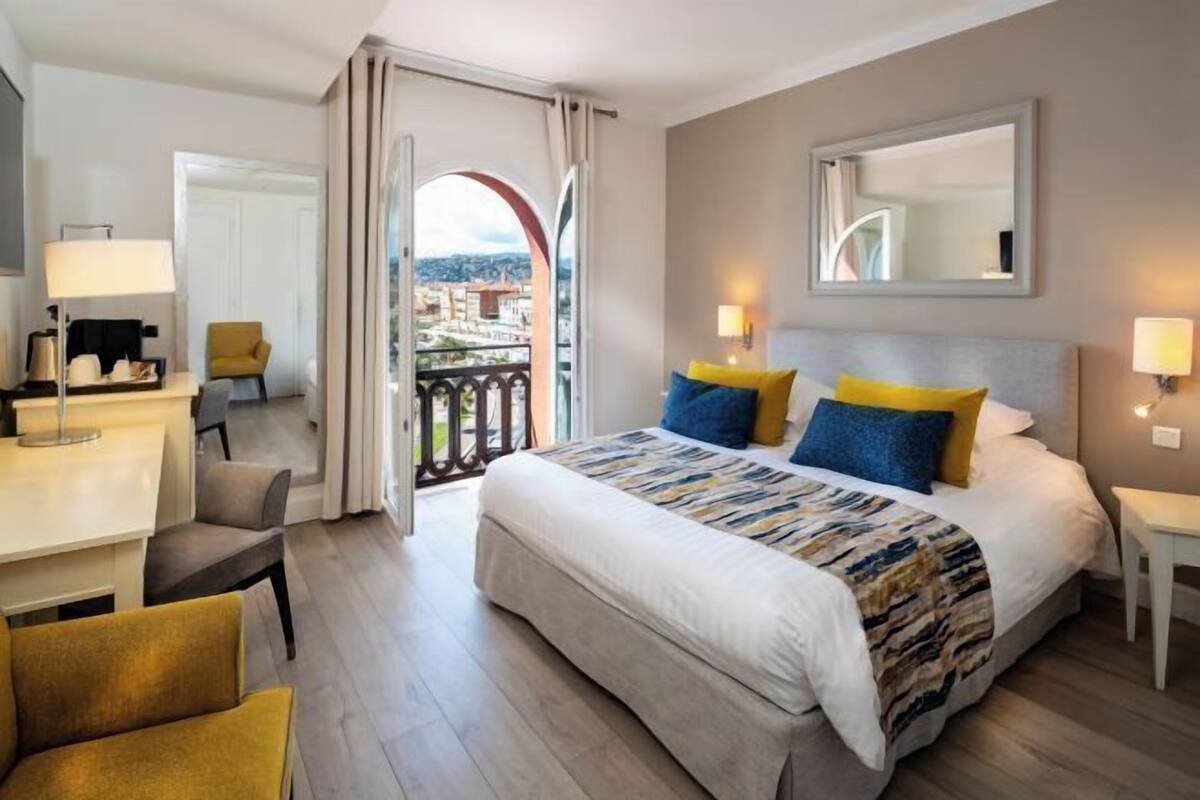 A hotel room with a bed, a desk, a chair, a balcony with a view, and a mirror on the wall.