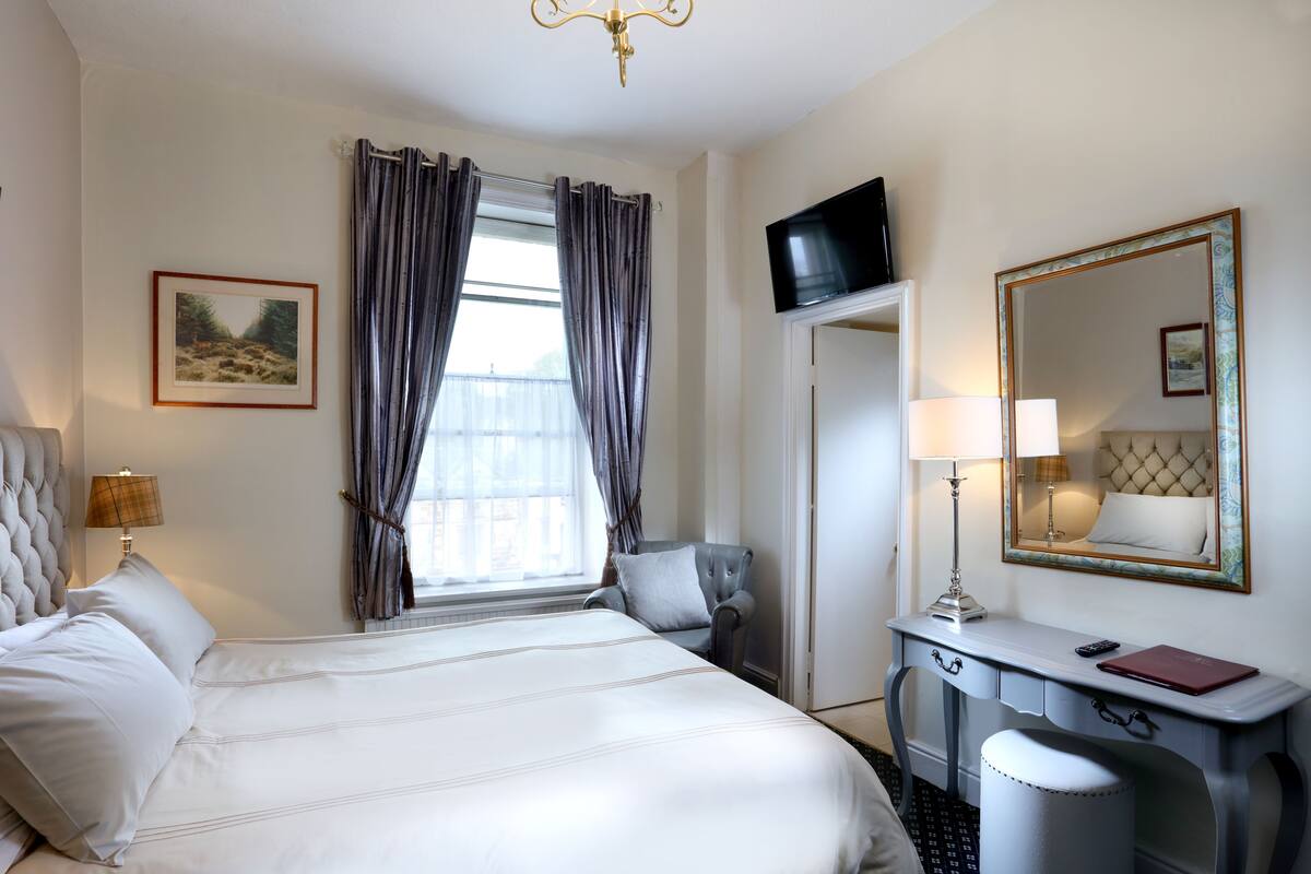 A hotel room with a large bed, a desk with a chair, a TV, a mirror, and a window with curtains.
