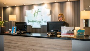 A Holiday Inn reception desk with a large sign, two computer monitors, a cash register, and a plant.