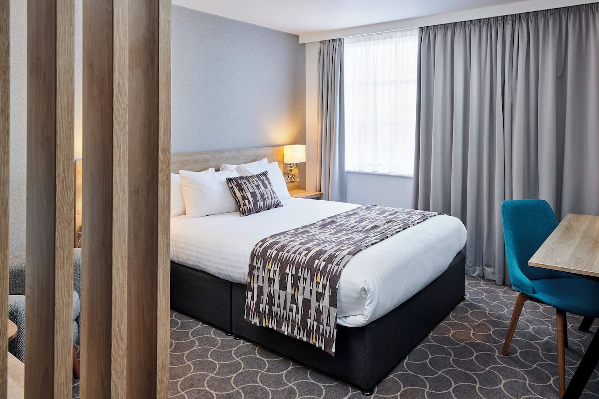 A hotel room with a large bed, a bedside table with a lamp, a chair, and a window with curtains.