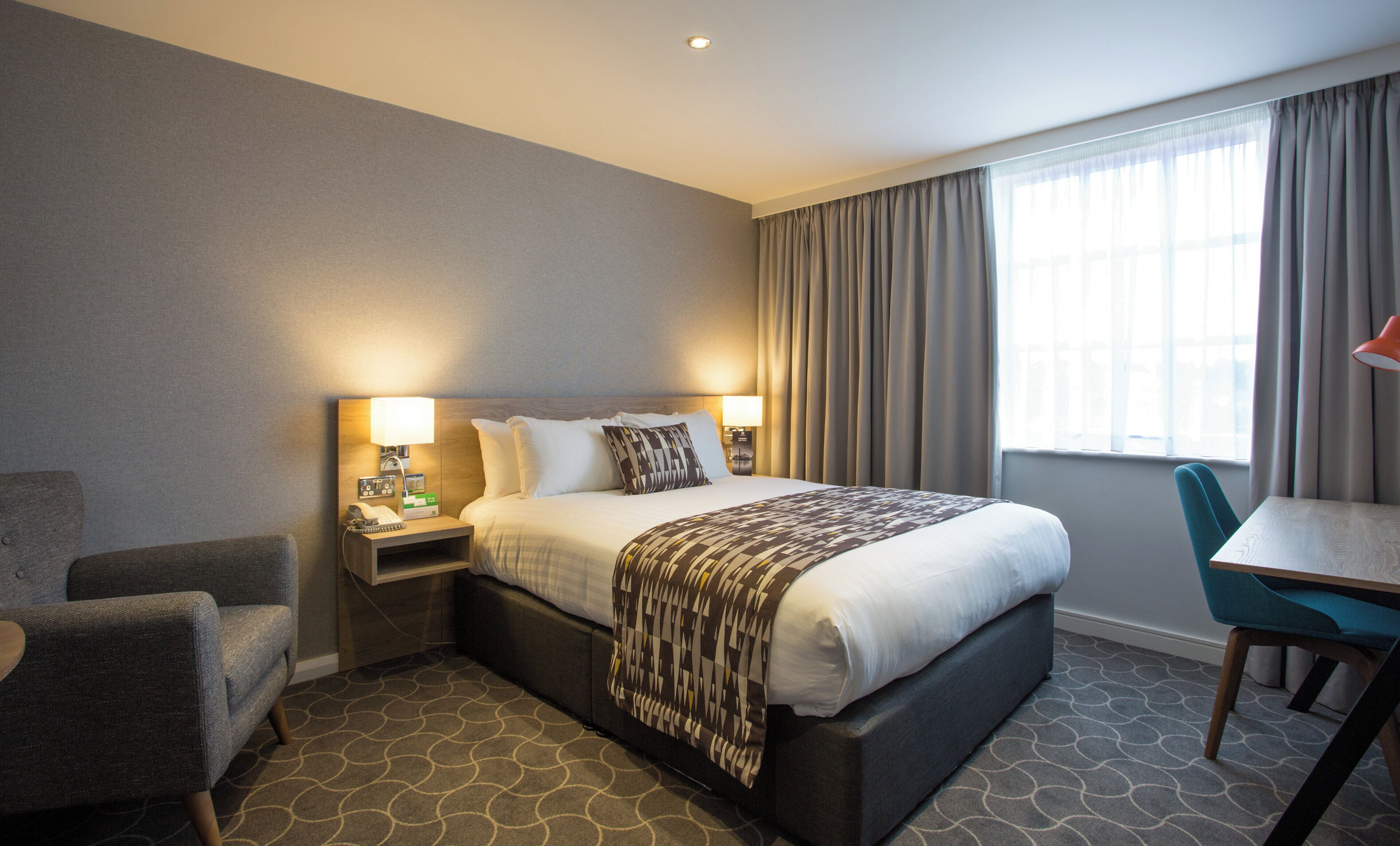 A hotel room with a bed, bedside tables, a desk, a chair, and a window with curtains.