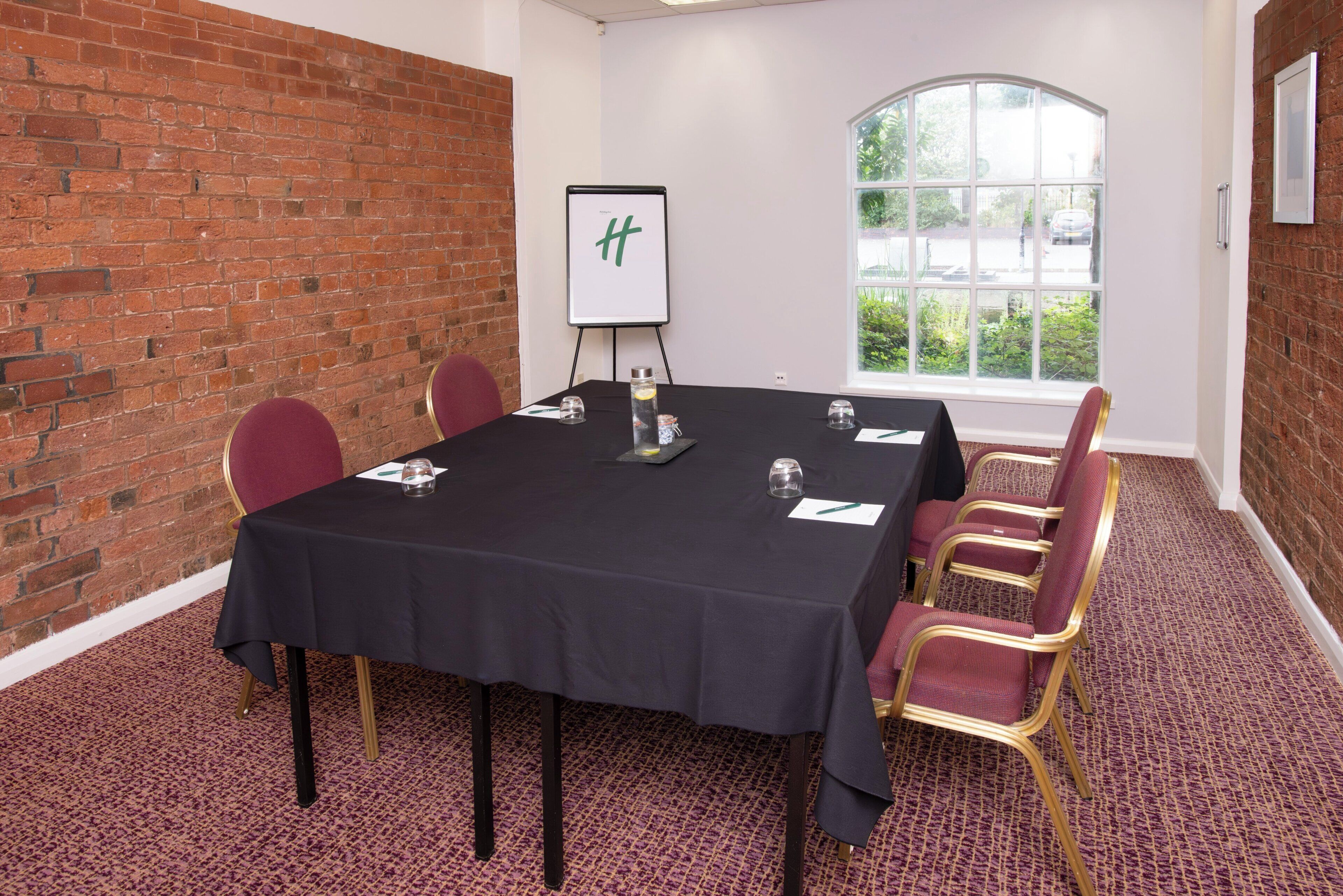 A conference room with a black table, red chairs, a whiteboard, and a window with a view of greenery.