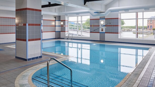 An indoor swimming pool with clear blue water, tiled walls, and a view of the outside through large windows.
