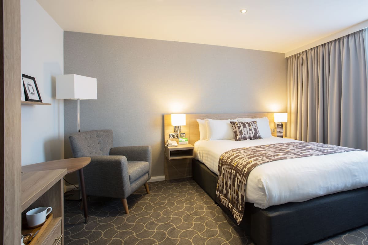 A hotel room with a bed, a chair, a desk, and a nightstand with lamps.