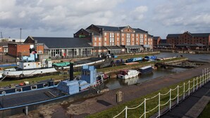 A riverside hotel with multiple boats docked, a pedestrian walkway, and a chain-link fence.