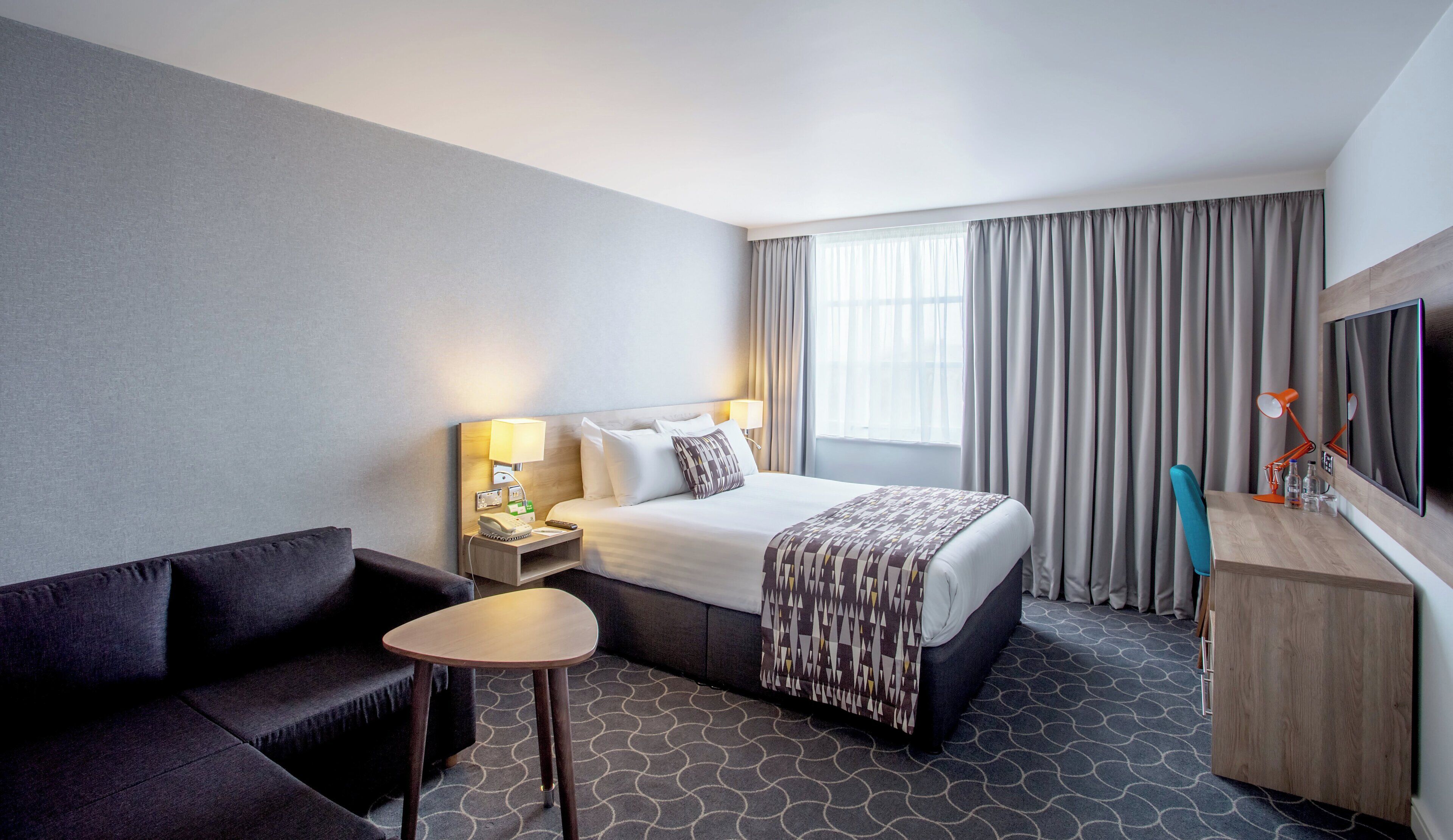 A hotel room with a bed, a desk, a chair, a TV, and a window with curtains.