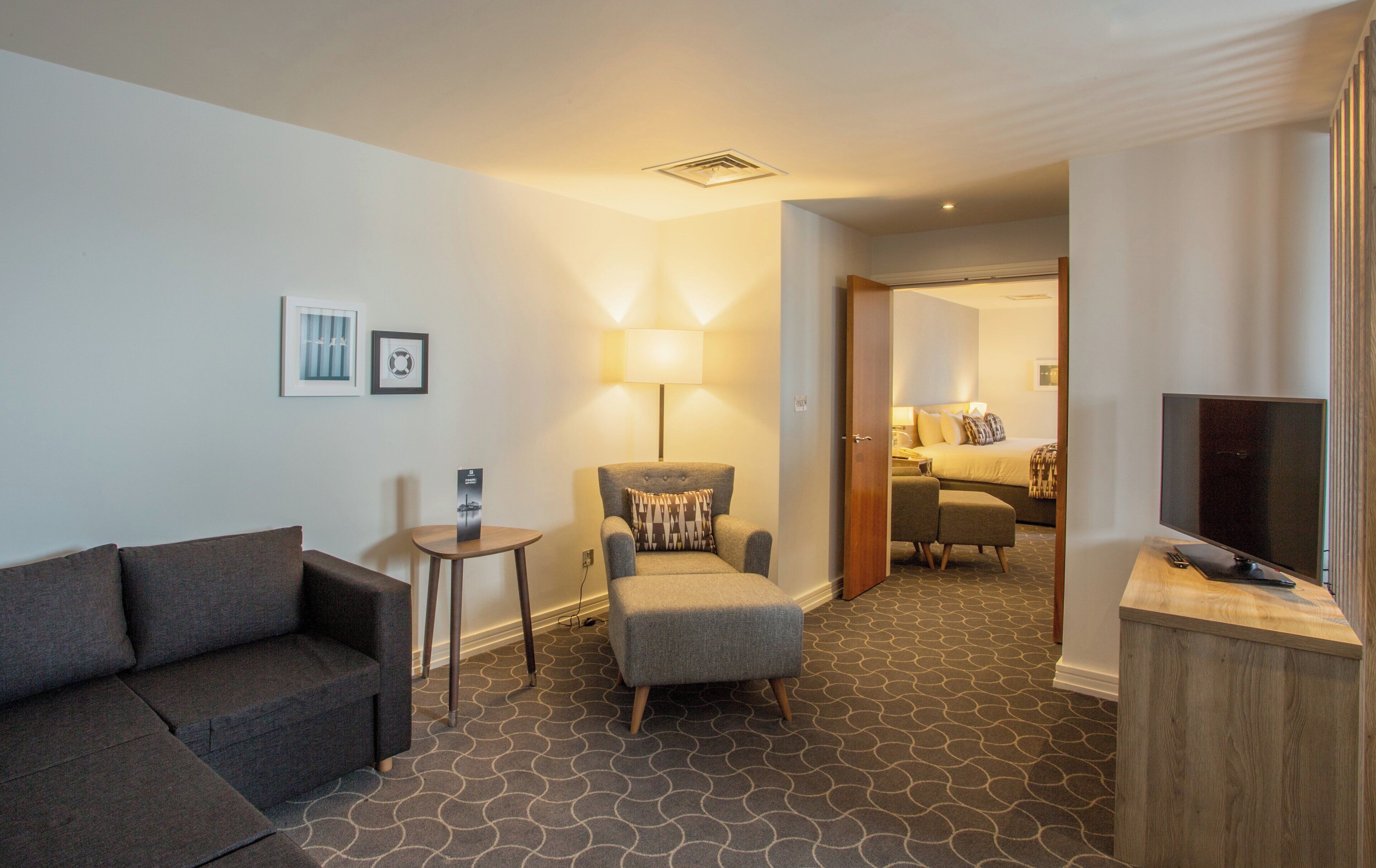 Een hotelkamer met een bank, fauteuil, klein tafeltje, lamp en een televisie op een houten standaard.