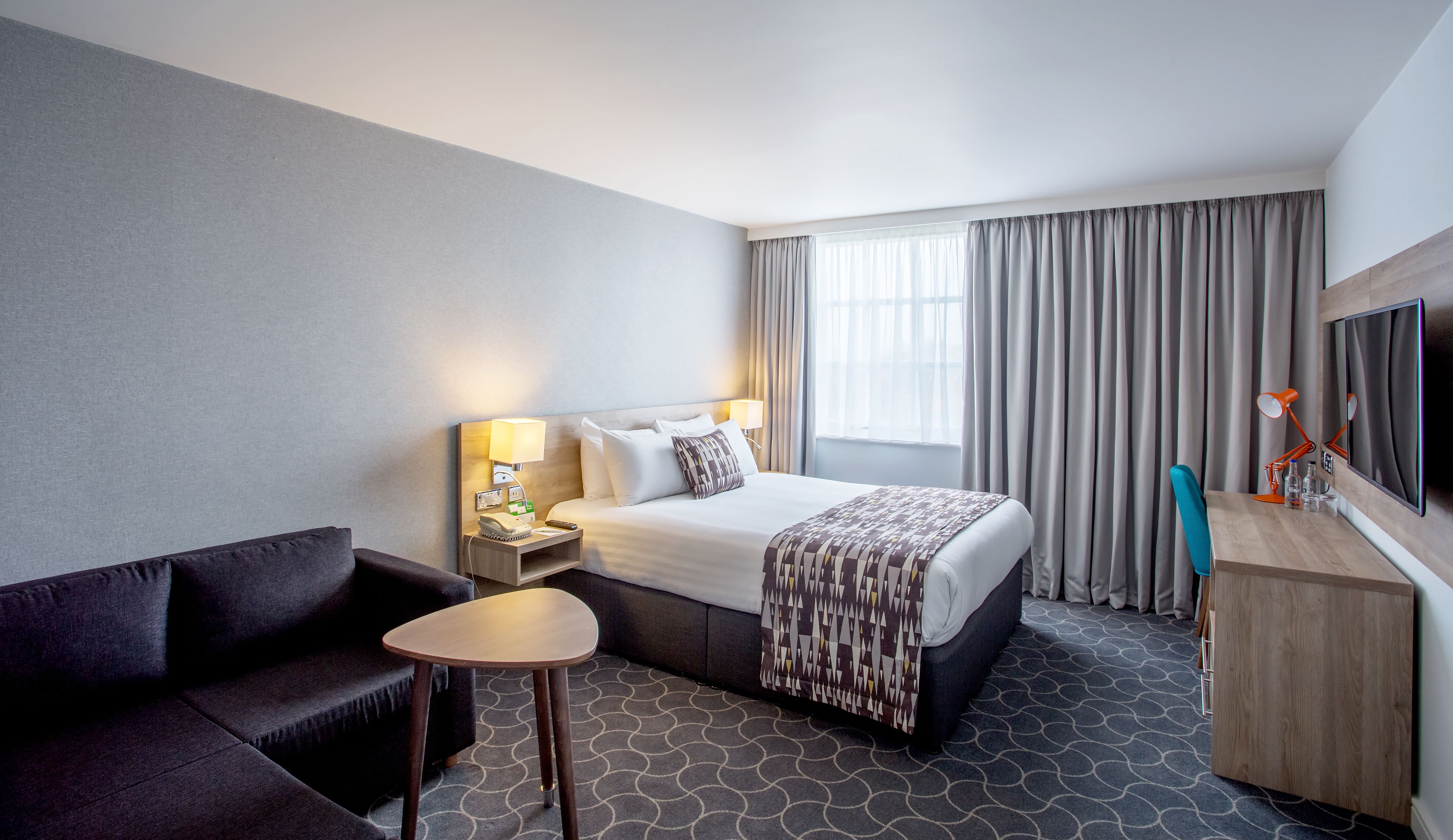 A hotel room with a bed, bedside table, lamp, TV, and a small seating area.