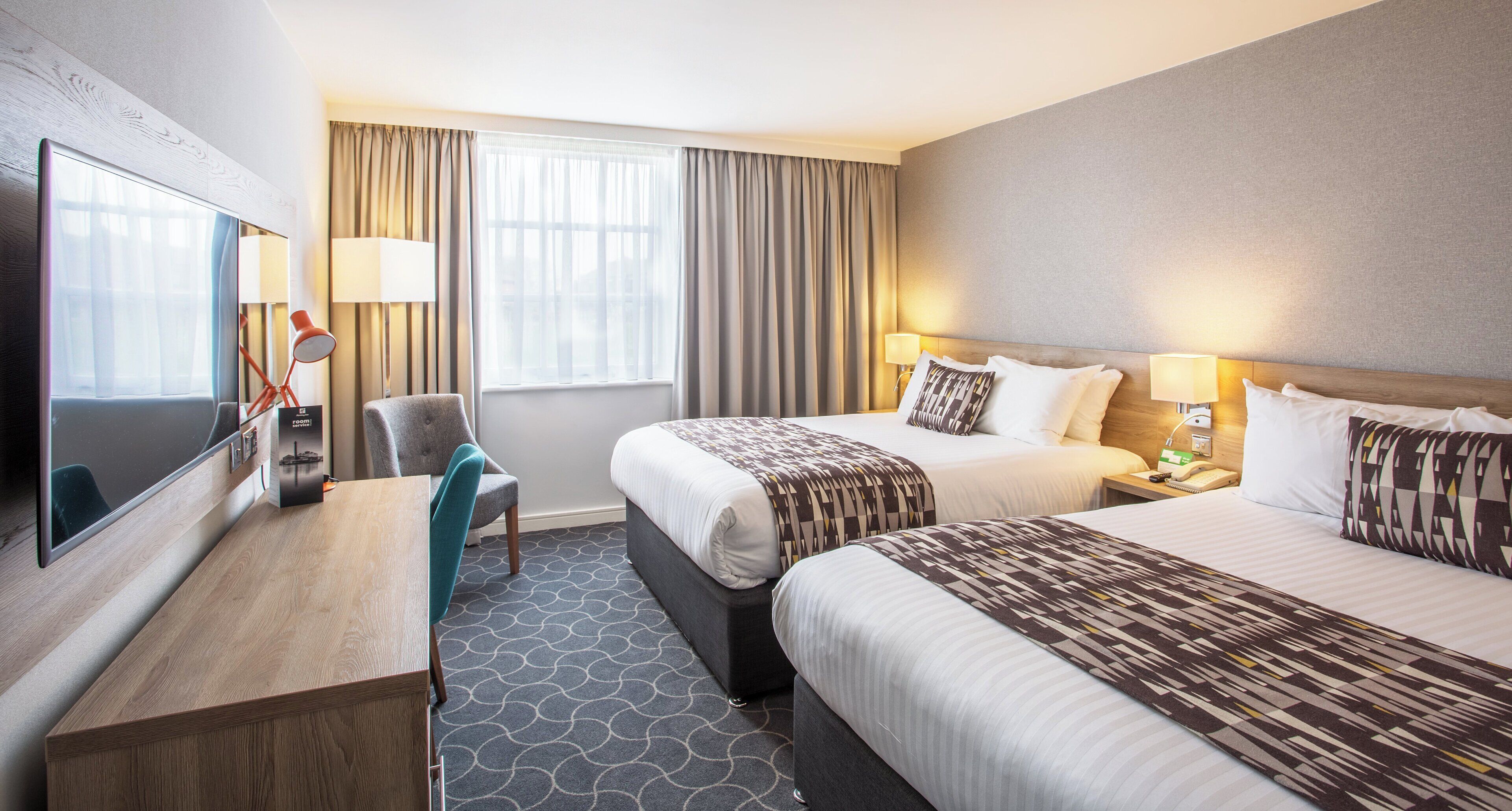 A hotel room with a bed, bedside tables, a desk, a chair, and a television.