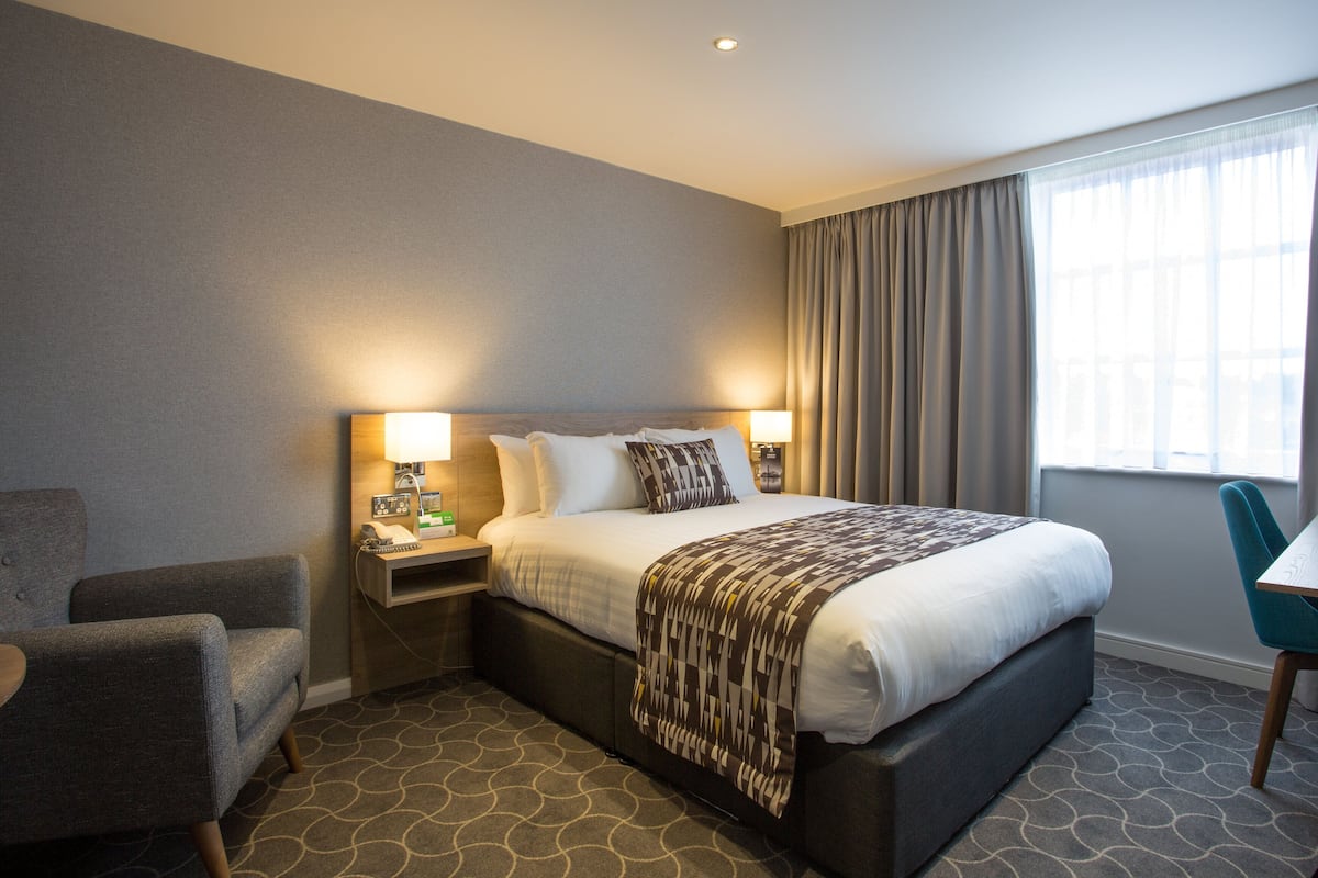 A hotel room with a bed, bedside tables, a desk, a chair, and a window with curtains.