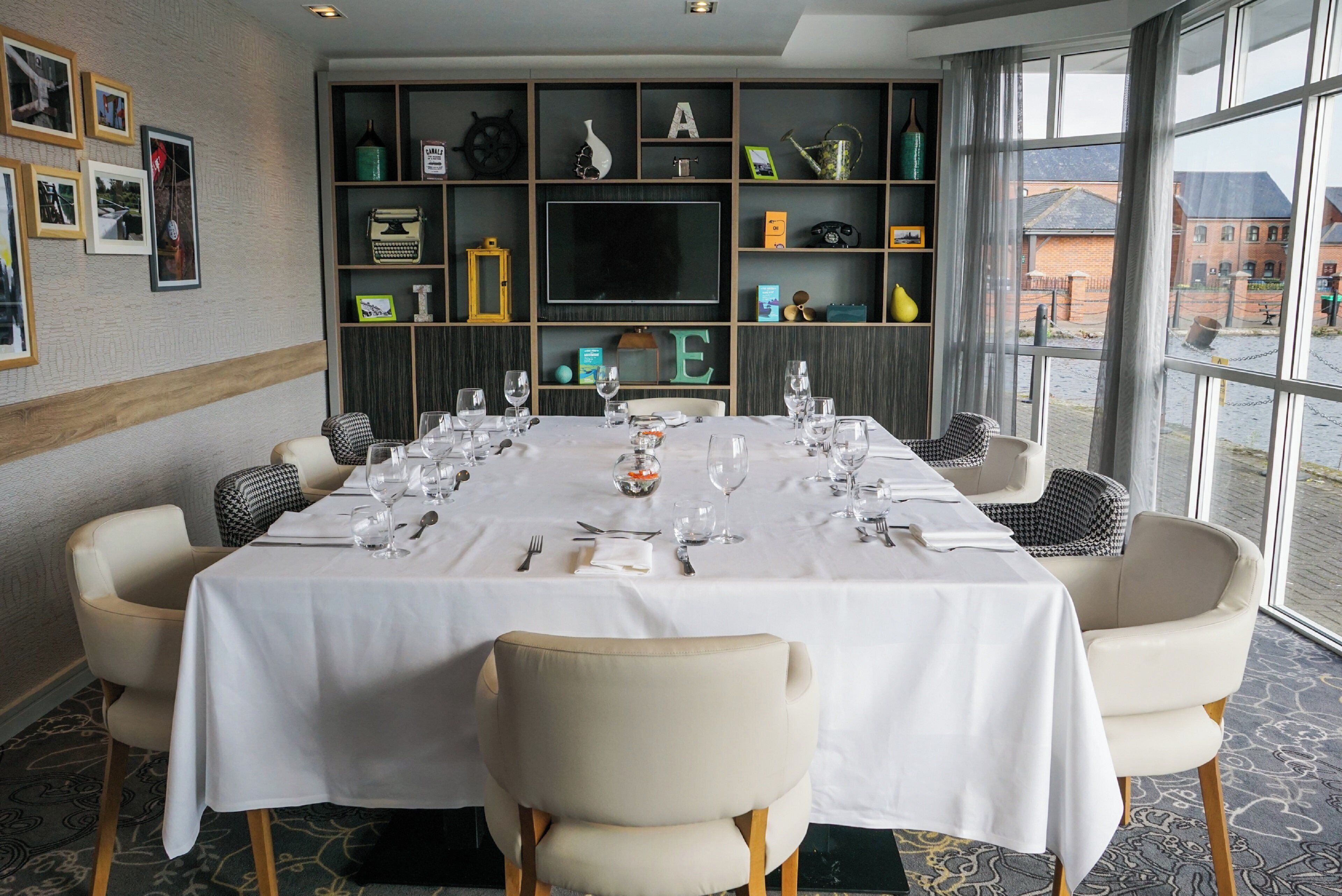 A dining area with a long table set for a meal, surrounded by chairs, with a bookshelf and decorative items in the background.
