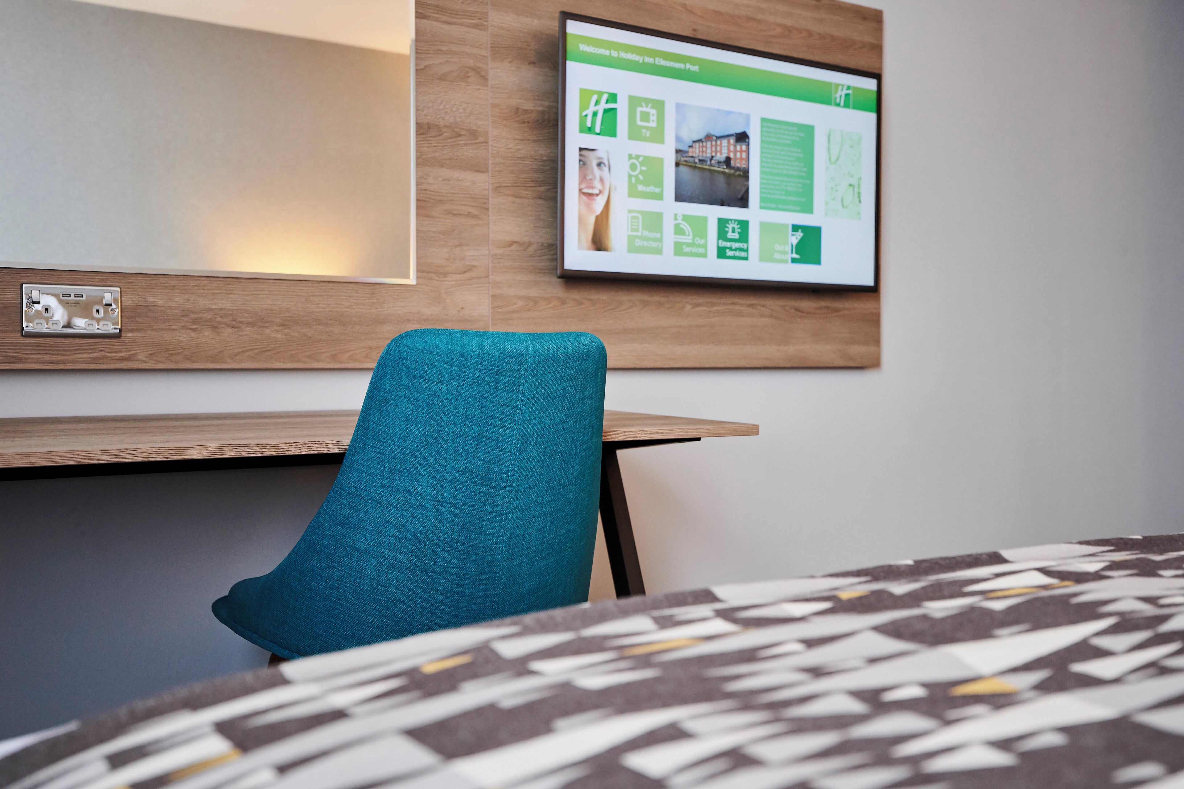 A hotel room with a wooden desk, a teal chair, and a framed informational poster on the wall.