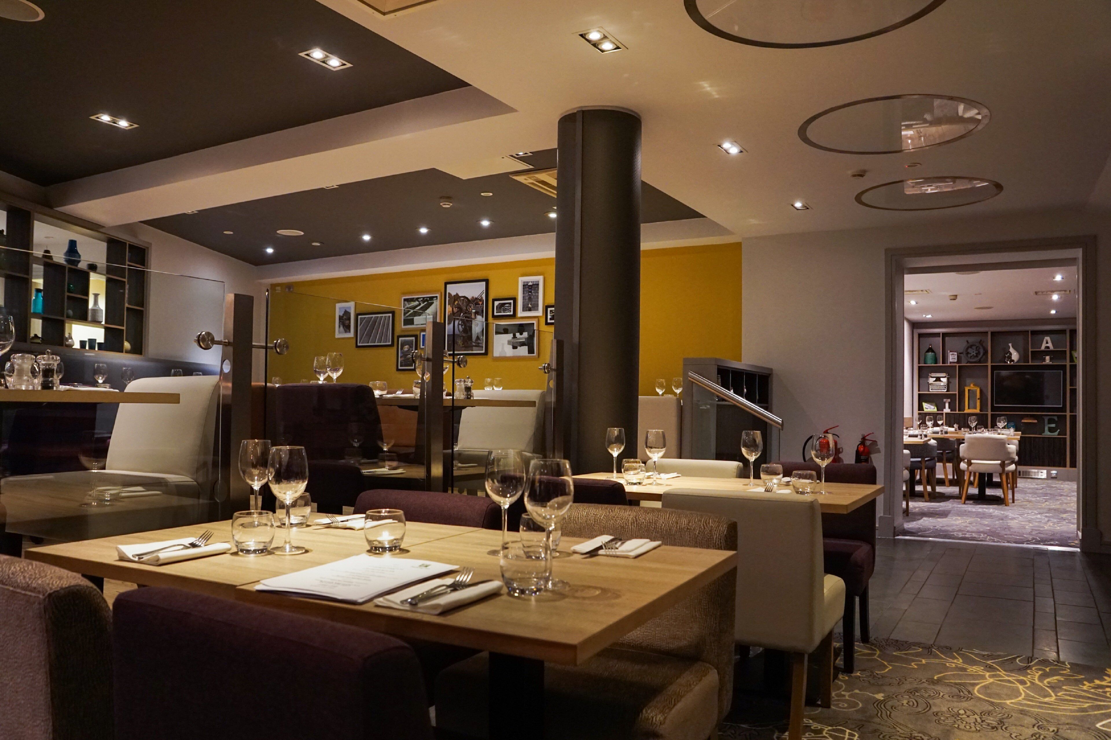 Un restaurante moderno con mesas dispuestas para comer, una zona de bar con vasos y botellas, y una pared de acento amarilla con cuadros enmarcados.