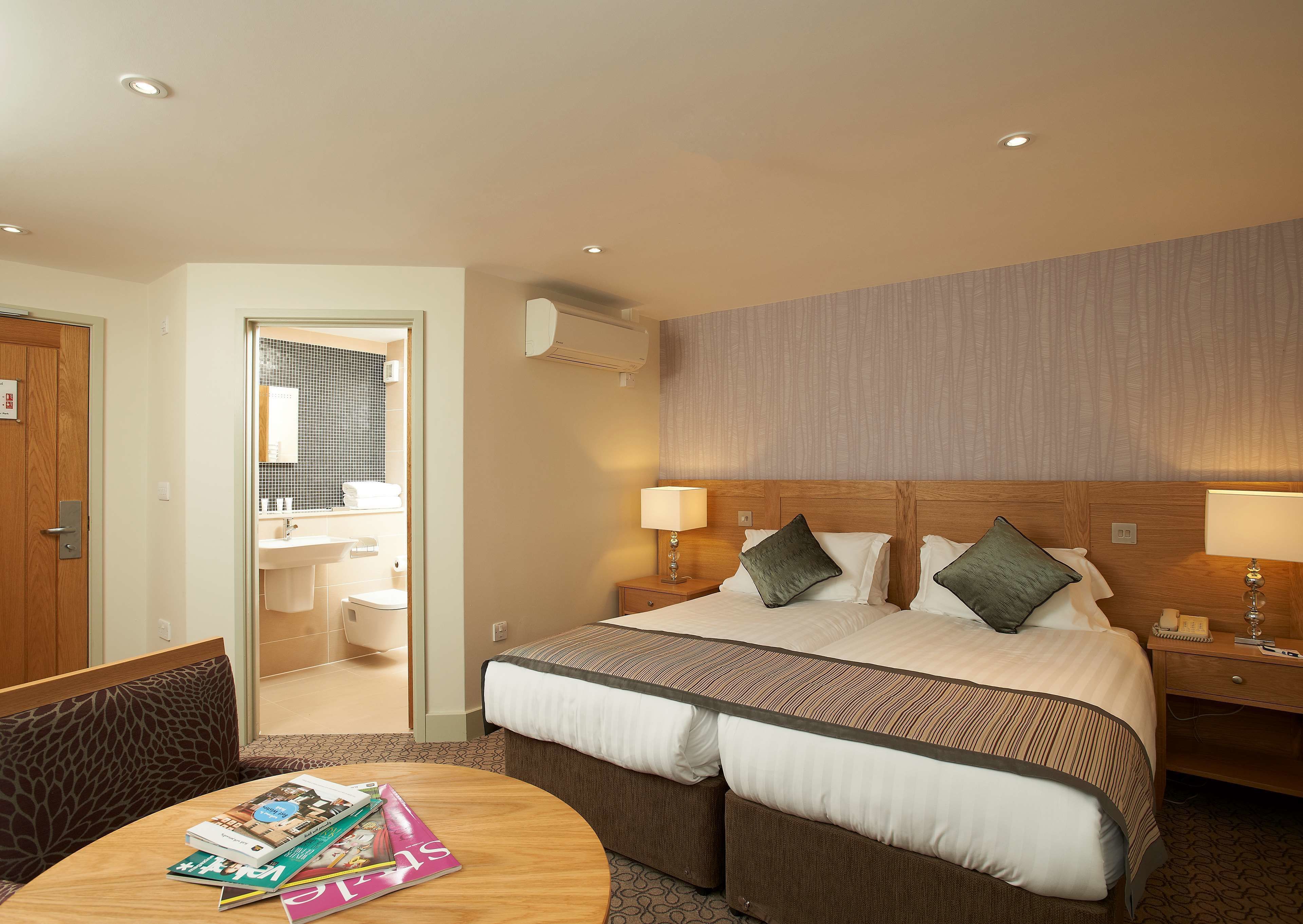 A hotel room with a large bed, two bedside tables with lamps, a wooden headboard, a small round table with magazines, and a bathroom visible through an open door.