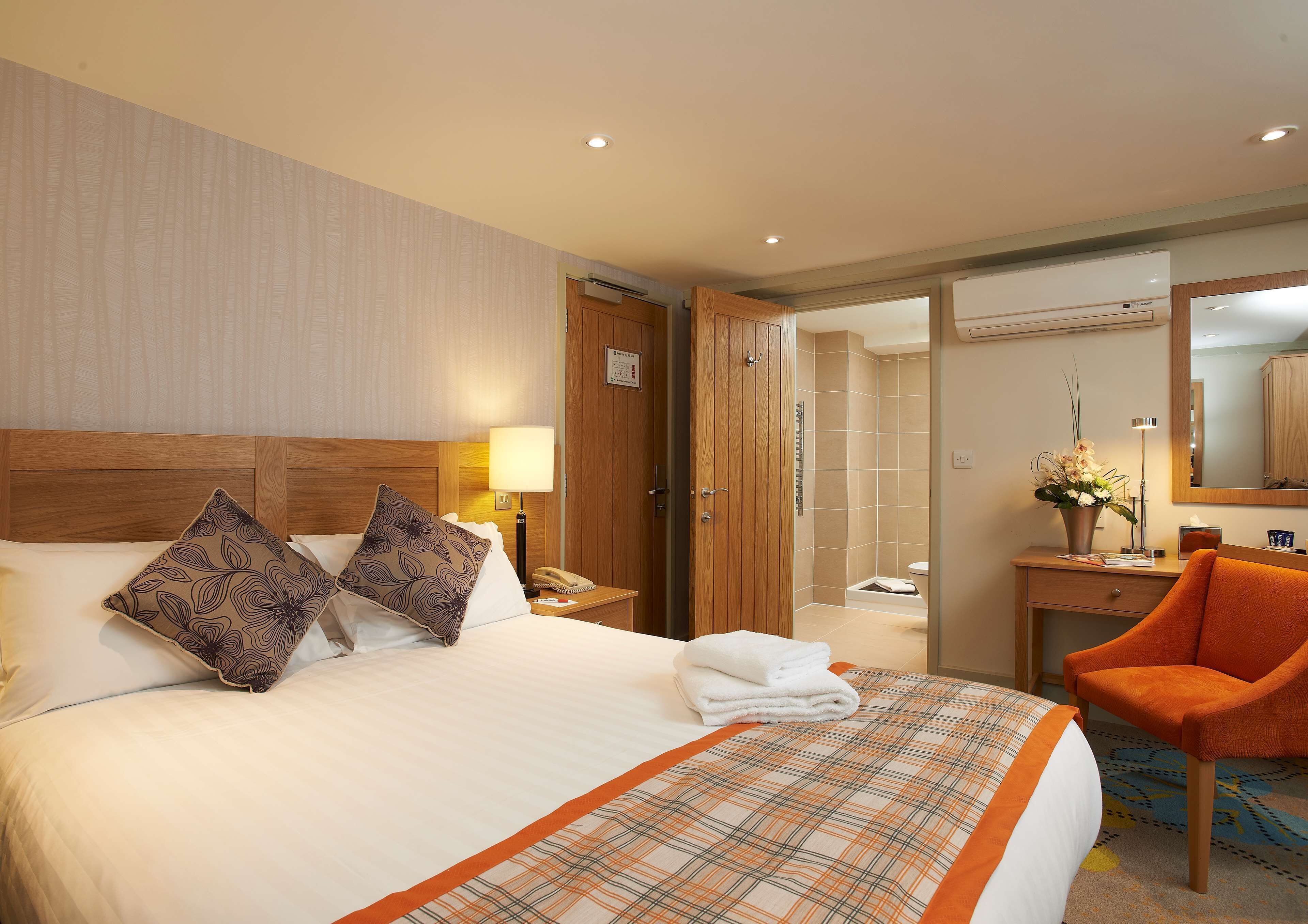 A hotel room with a large bed, two decorative pillows, a bedside lamp, a wooden nightstand, a bathroom visible through an open door, a desk with a chair, and a mirror.