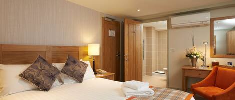 A hotel room with a large bed, two decorative pillows, a bedside lamp, a wooden nightstand, a bathroom visible through an open door, a desk with a chair, and a mirror.