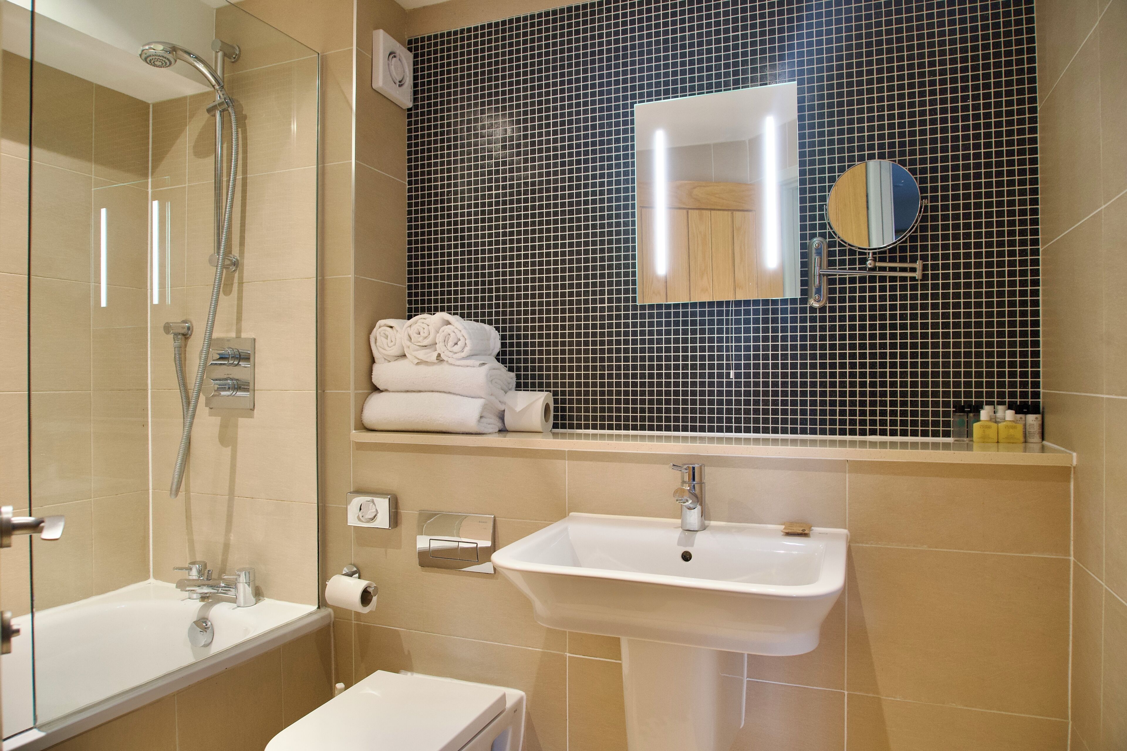 A bathroom with a shower, a sink, a mirror, and neatly rolled towels.