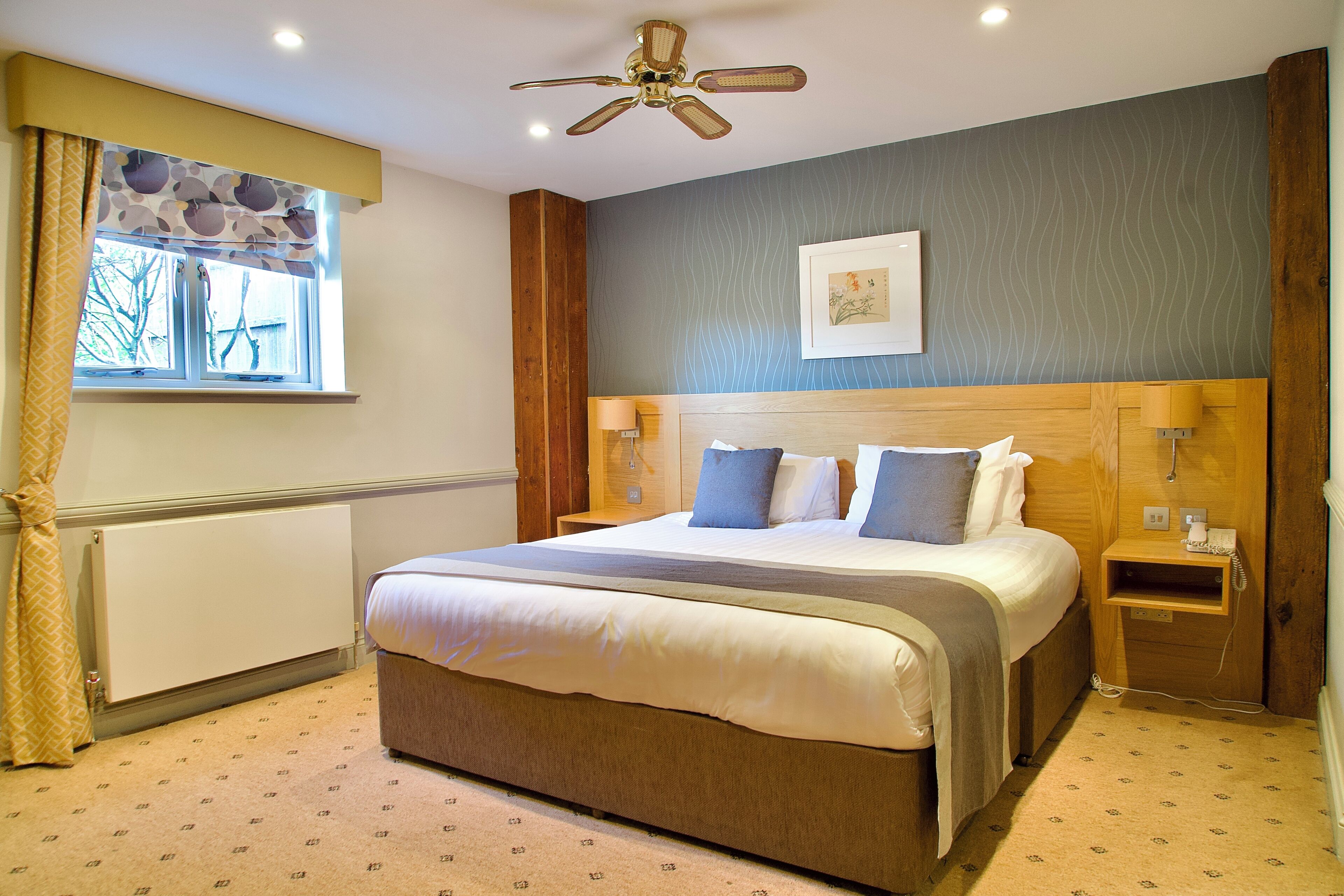 A hotel room with a bed, a ceiling fan, a window with curtains, a bedside table, and a wall-mounted picture.