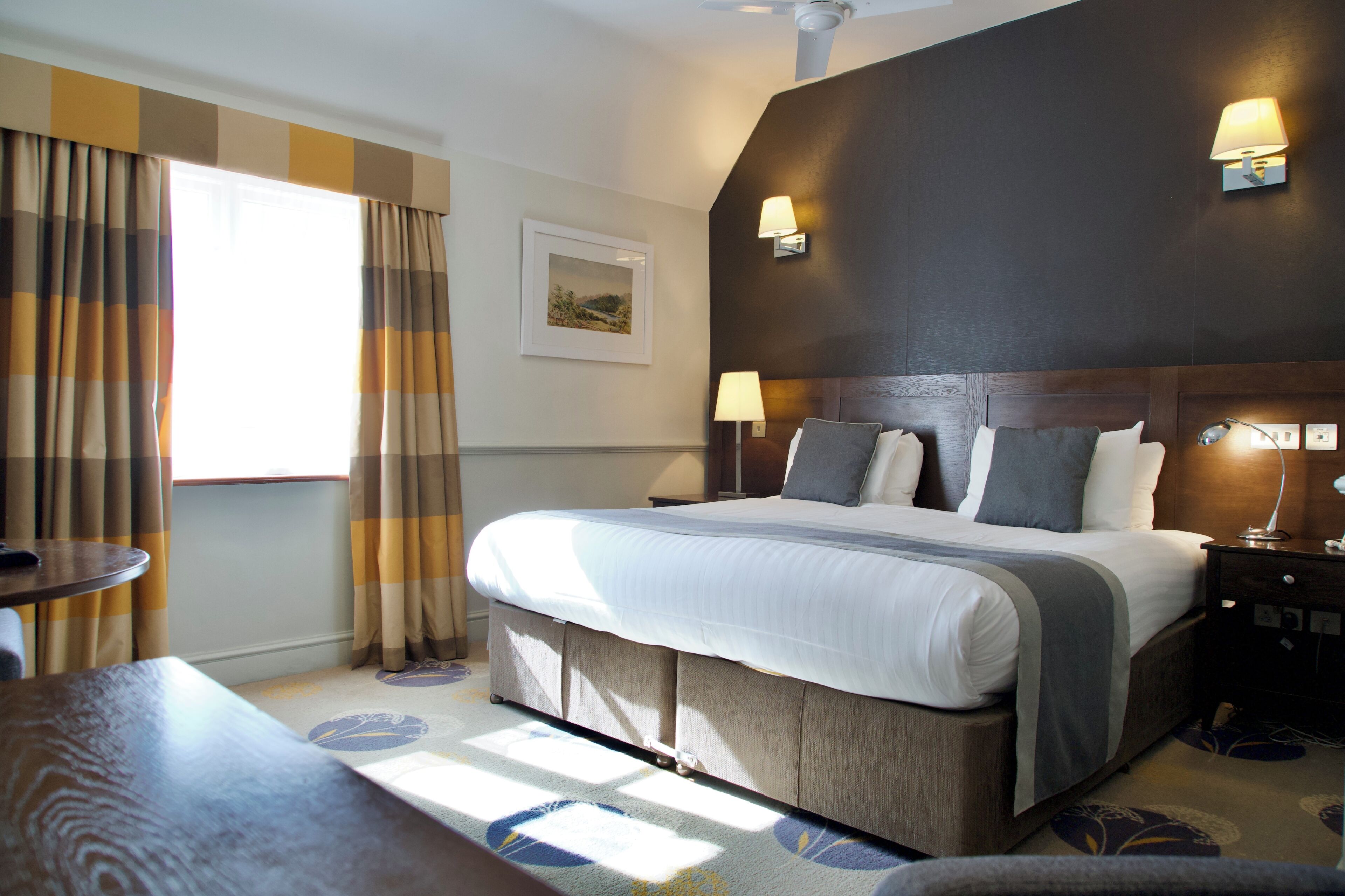 Habitación de hotel con cama, lámparas de noche, mesita de noche, mesa y ventana con cortinas.