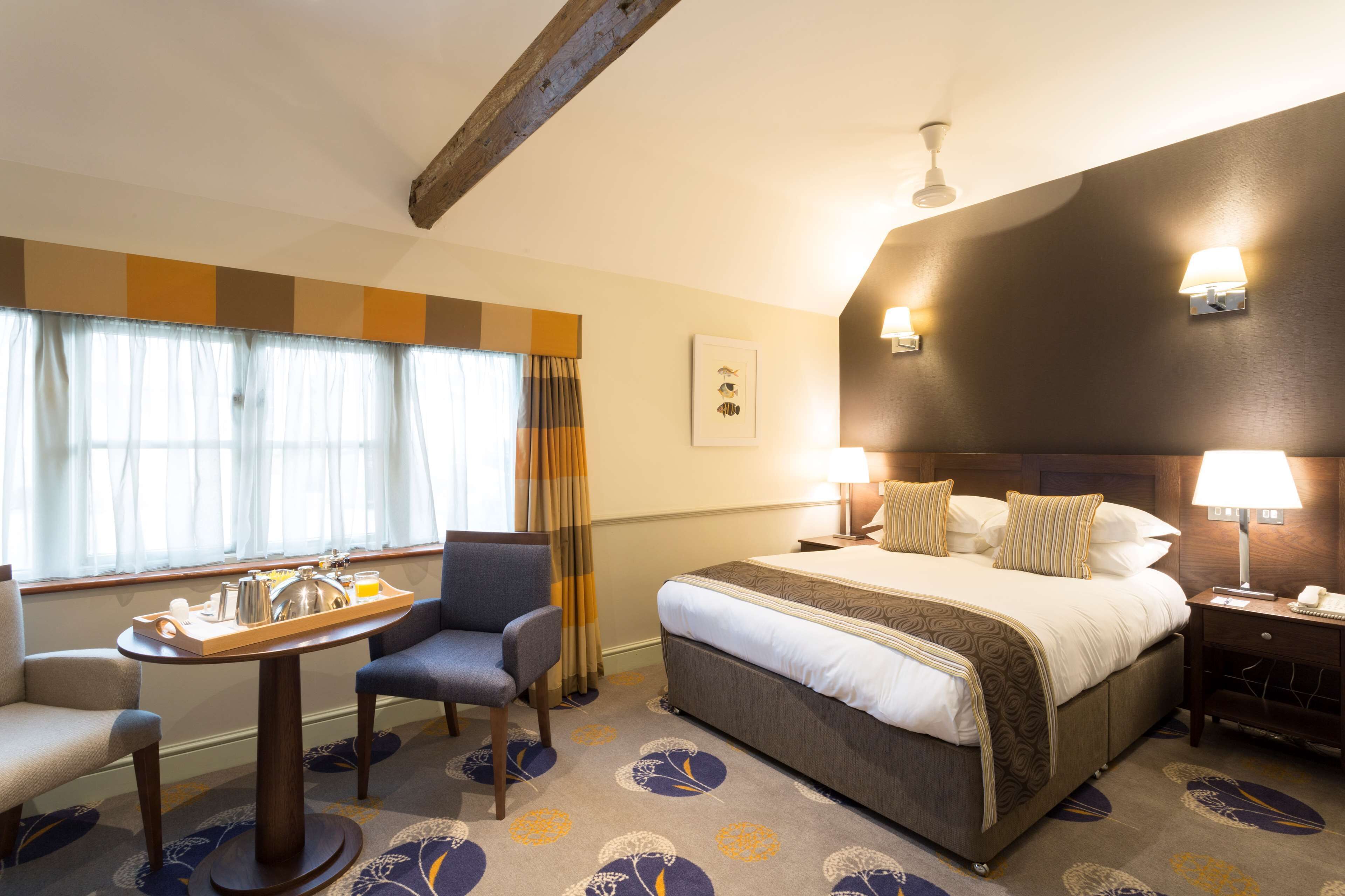 Una habitación de hotel con una cama, un escritorio con una bandeja de artículos de desayuno, una silla y una mesita de noche con una lámpara.