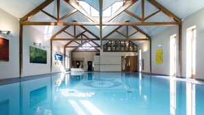 Une piscine intérieure dotée d’un plafond haut et de poutres en bois, entourée de murs ornés d’œuvres d’art et de fenêtres.