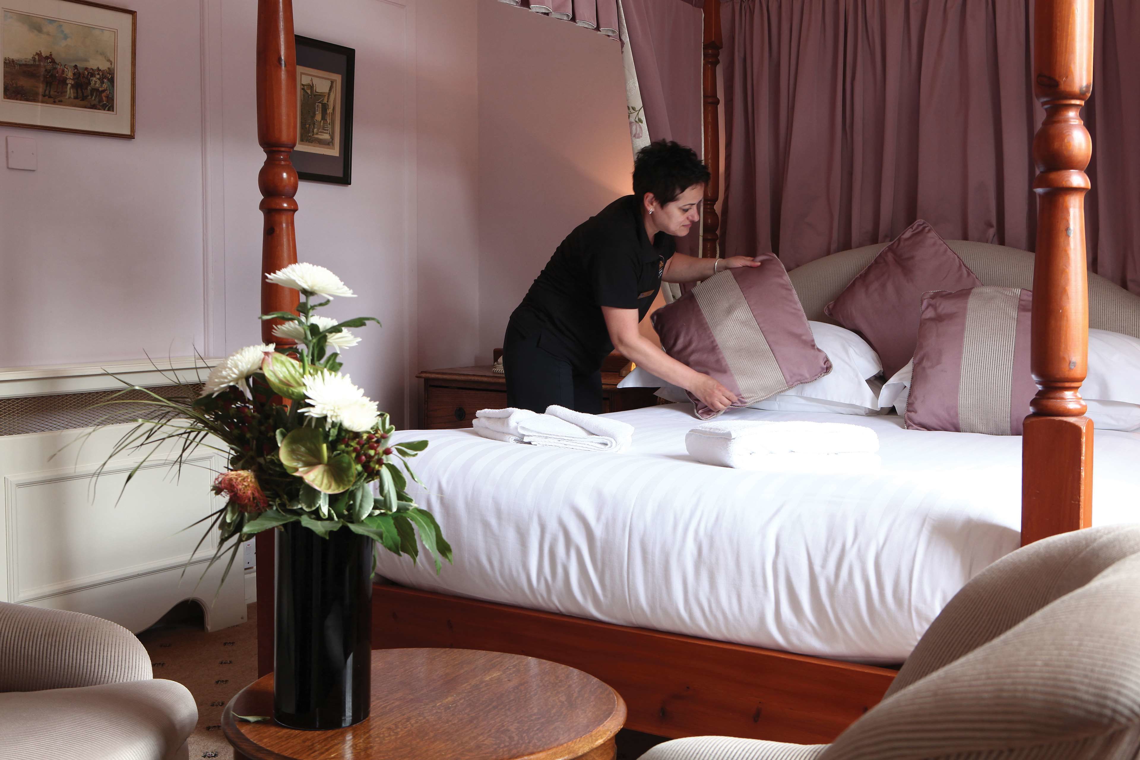 A neatly made bed with white linens and multiple pillows, a vase with flowers on a wooden table, and a wooden headboard.