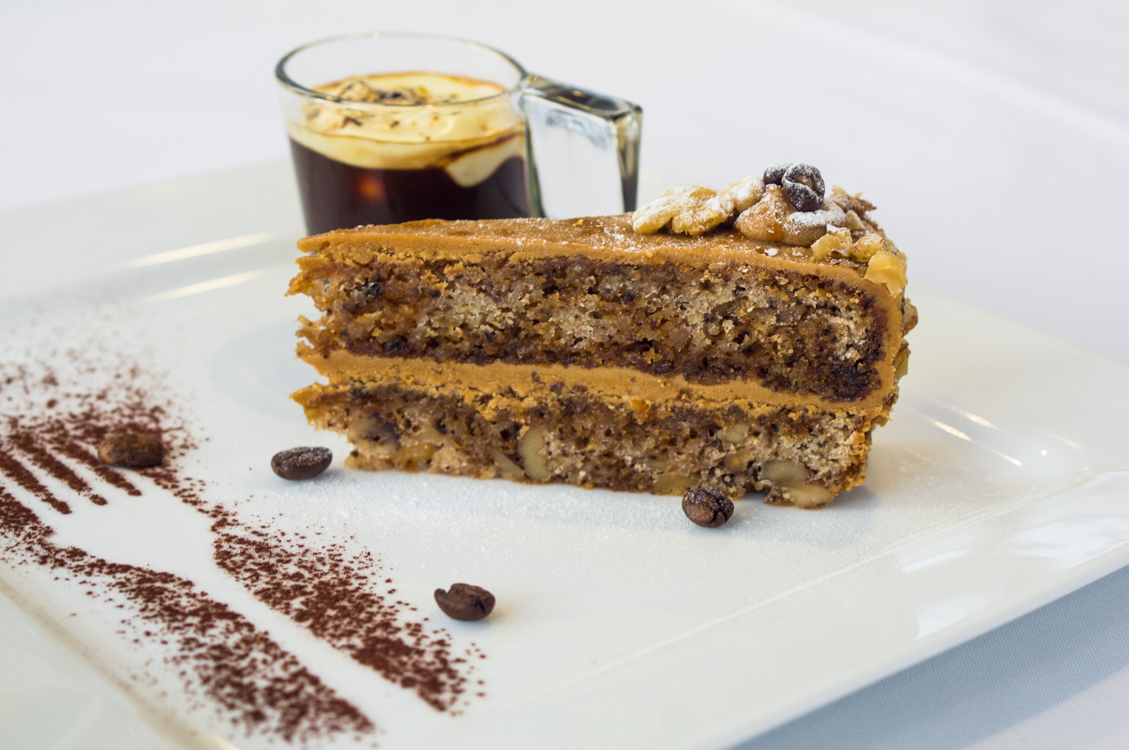 Une part de gâteau à plusieurs couches, accompagnée d’un verre de café, sur une assiette blanche.