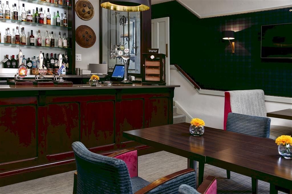 A bar area with a wooden counter, shelves of liquor bottles, a beer tap, and a digital screen. There is a dining area with a wooden table and chairs, and a green accent wall.