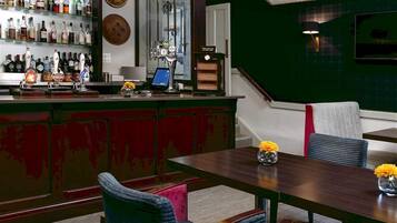 A bar area with a wooden counter, shelves of liquor bottles, a beer tap, and a digital screen. There is a dining area with a wooden table and chairs, and a green accent wall.