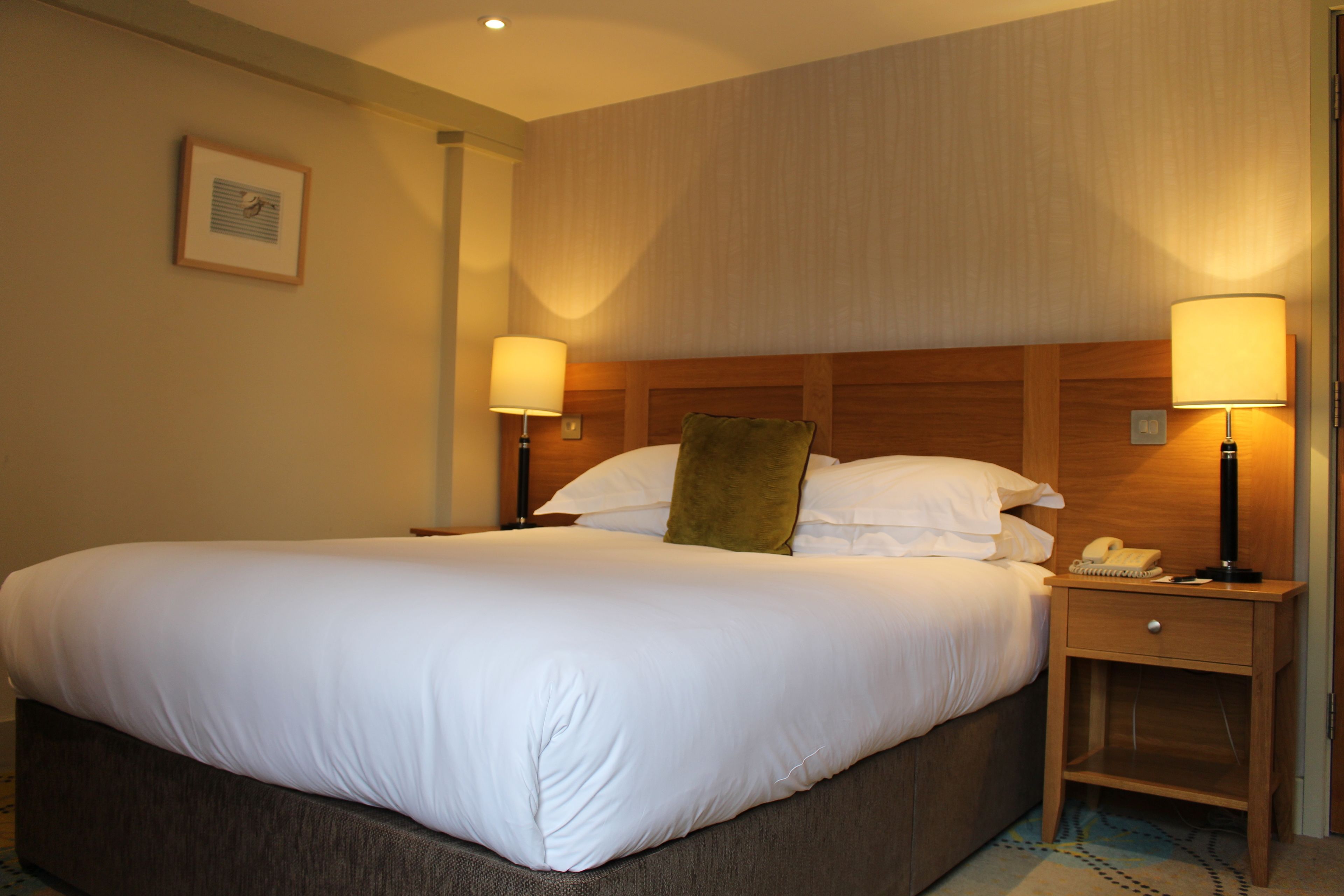 A hotel room with a large bed, two bedside tables with lamps, a wooden headboard, and a framed picture on the wall.