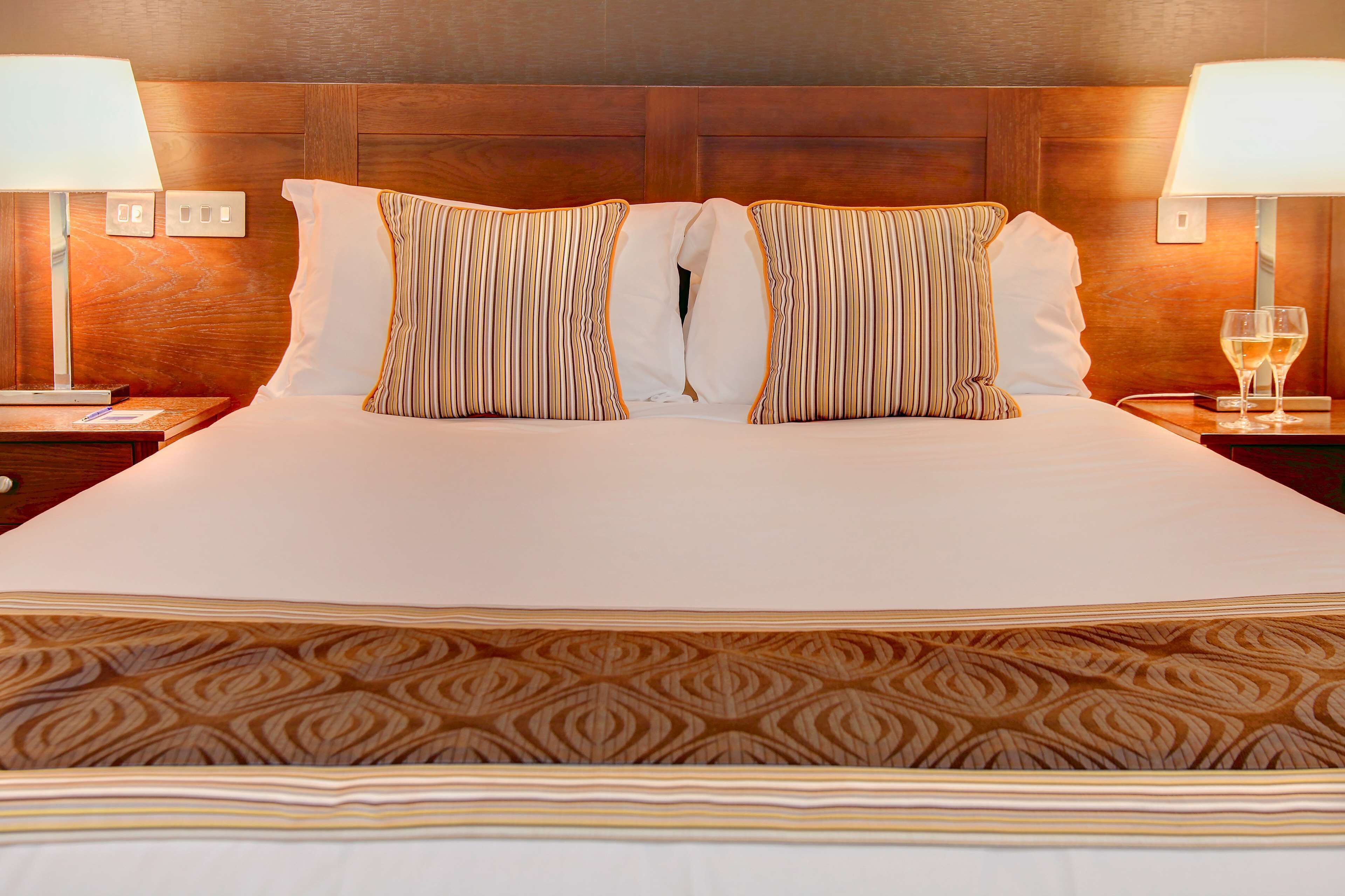 A neatly made bed with striped pillows, a patterned bedspread, and two bedside lamps with glasses of wine on the nightstand.