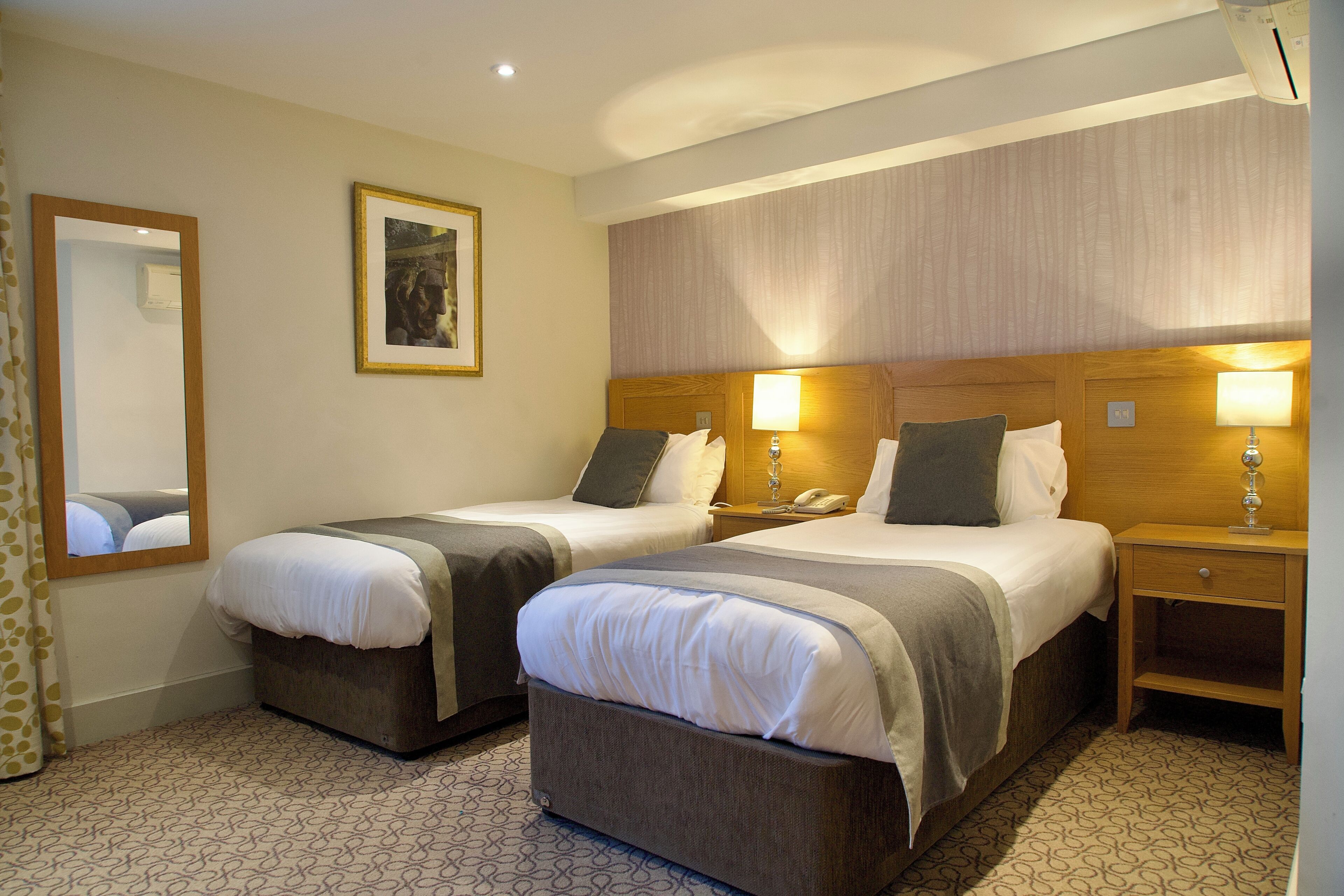 Une chambre d’hôtel avec deux lits, une table de chevet avec une lampe, un miroir et un tableau encadré au mur.