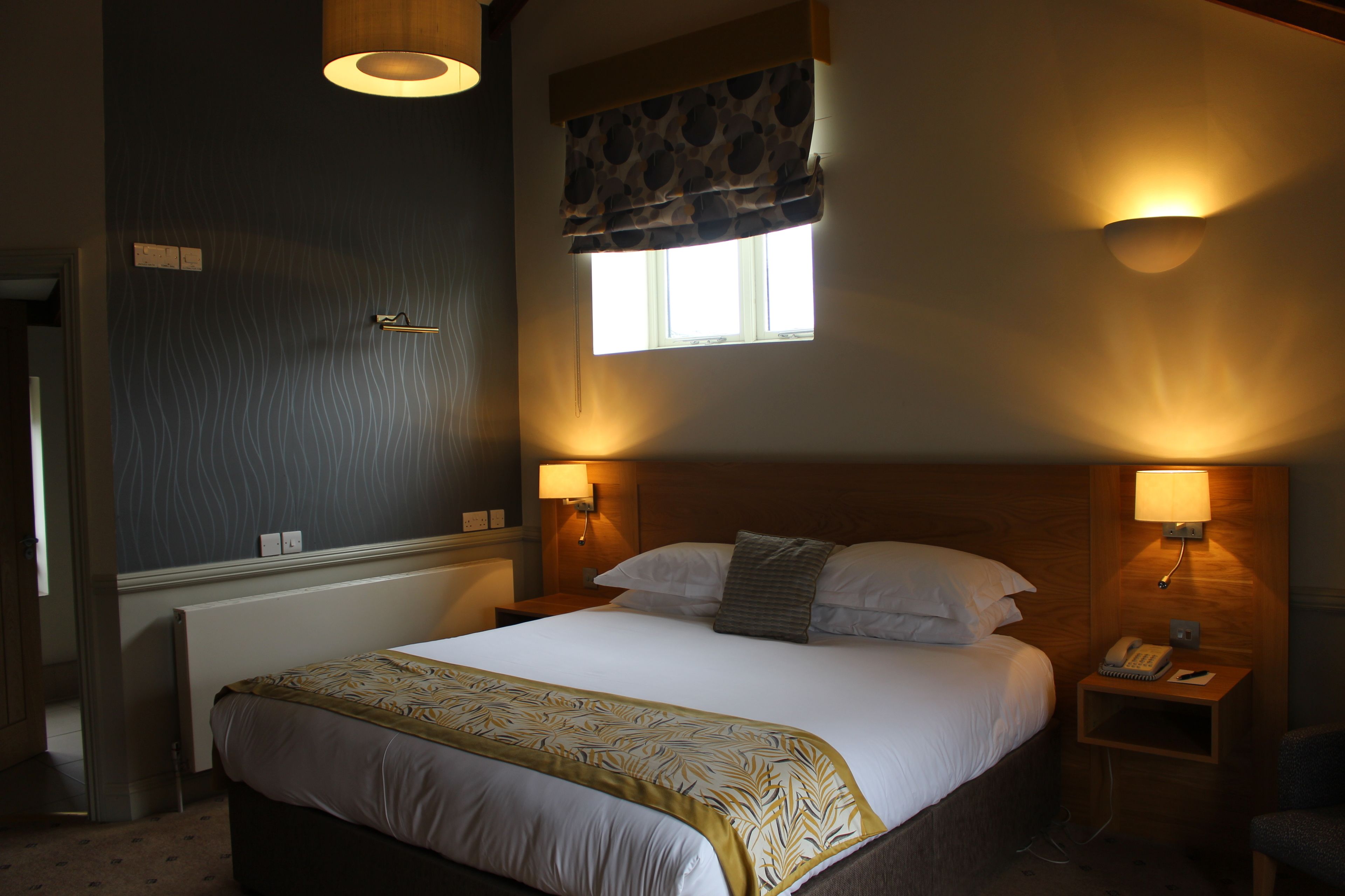 A neatly made bed with a patterned bedspread, two bedside lamps, a nightstand with a phone, and a window with a patterned curtain.
