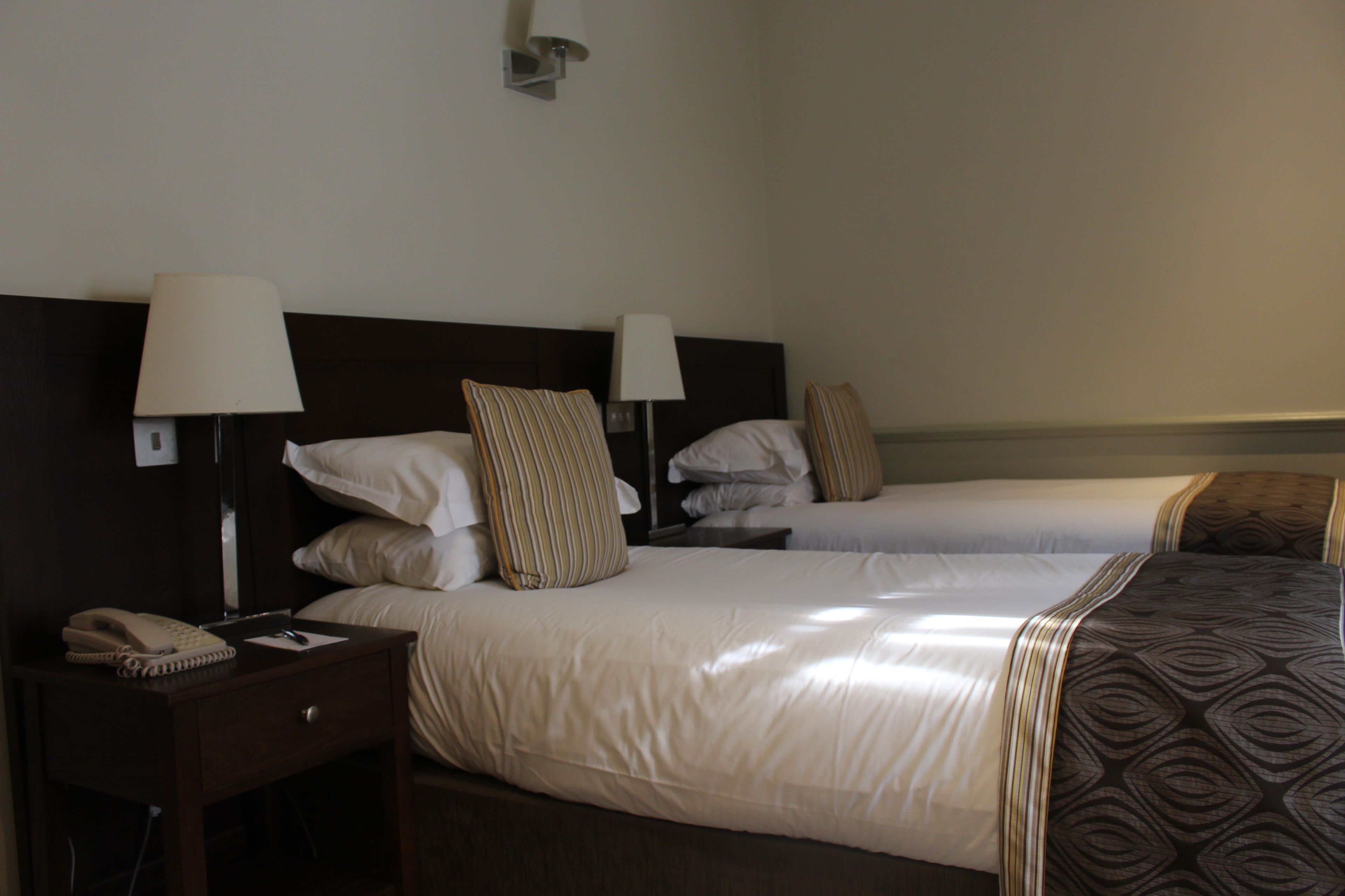 A hotel room with two beds, each with a pillow and a lamp. There is a nightstand with a phone and a lamp.