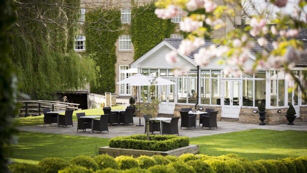 Un espace extérieur aménagé en terrasse, avec des tables et des chaises, entouré de haies bien entretenues et bordé d’un bâtiment doté de grandes fenêtres.