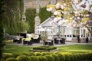 An outdoor patio area with tables and chairs, surrounded by well-maintained hedges and a building with large windows. - Cambridge Quy Mill Hotel & Spa in Cambridge (Cambridge)