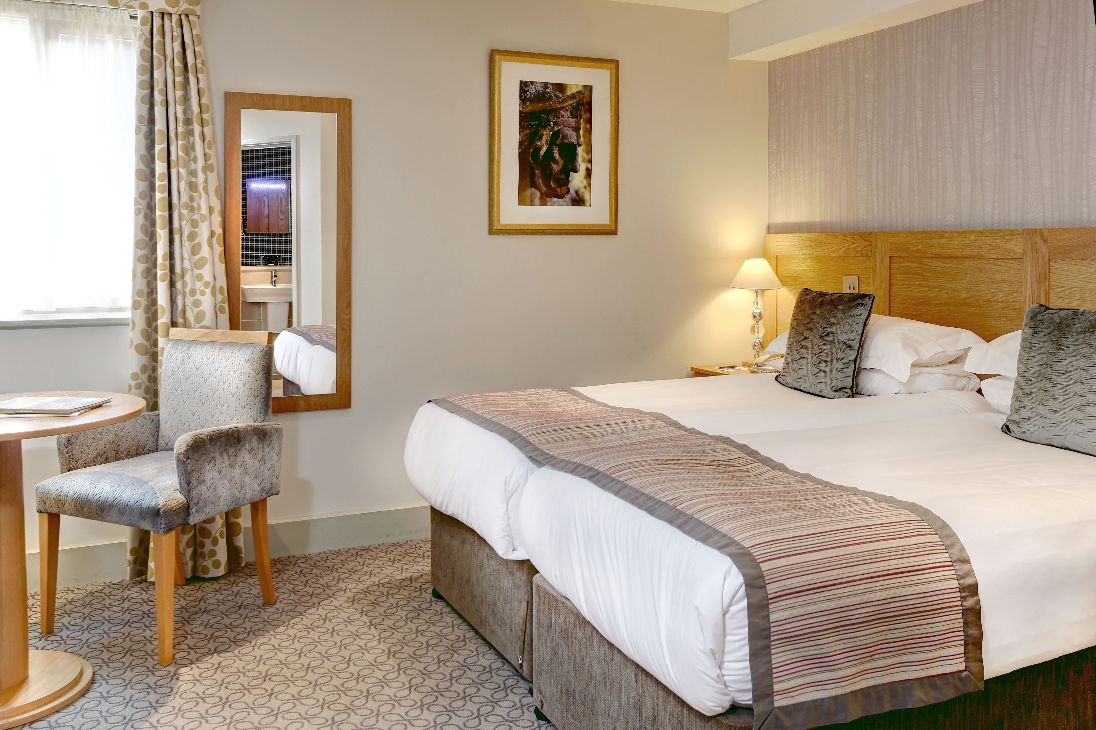 A hotel room with a bed, a chair, a table, a mirror, and a picture on the wall.