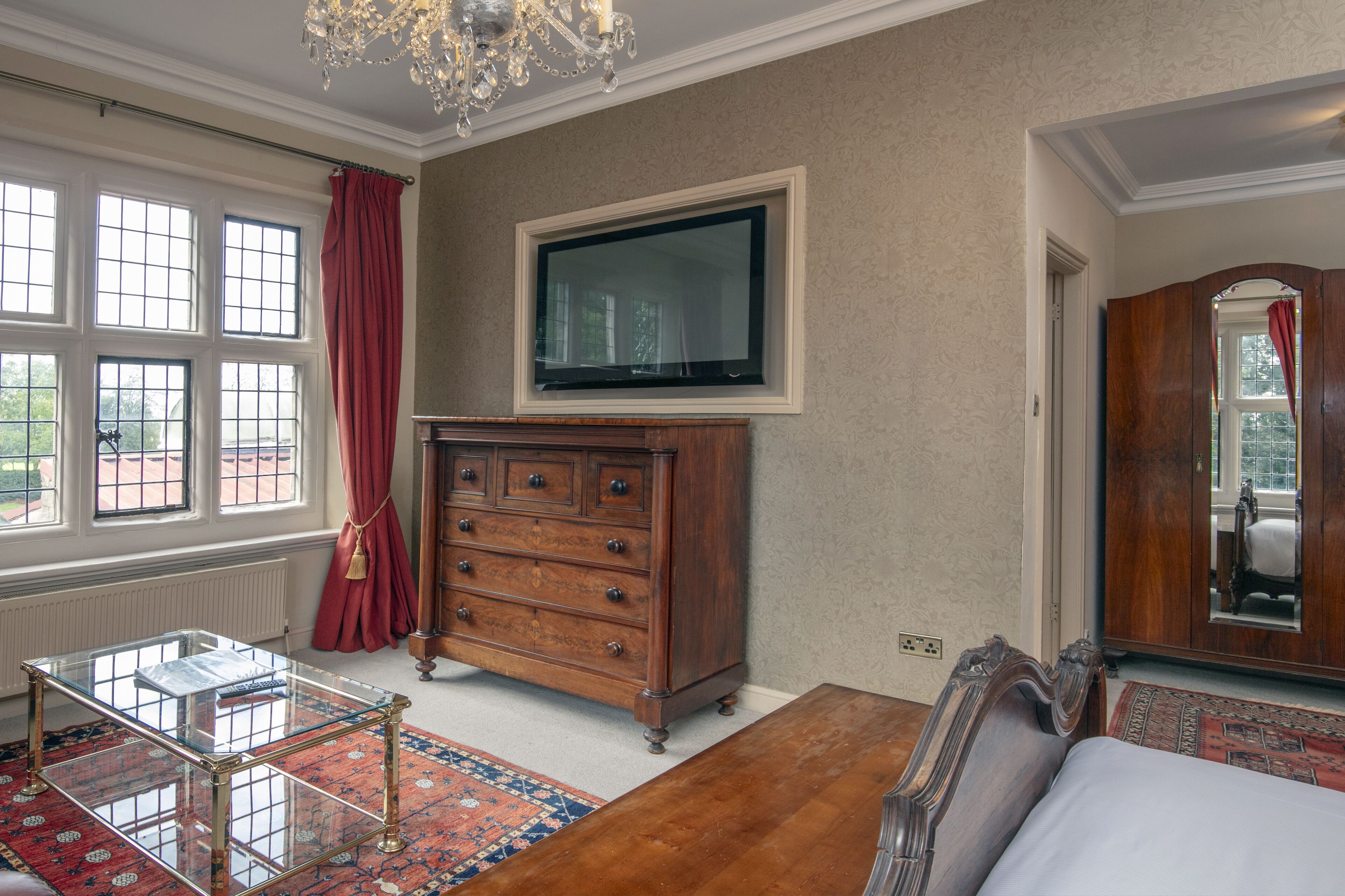 A room with a wooden dresser, a glass coffee table, a bed, a television mounted on the wall, and a large window with red curtains.