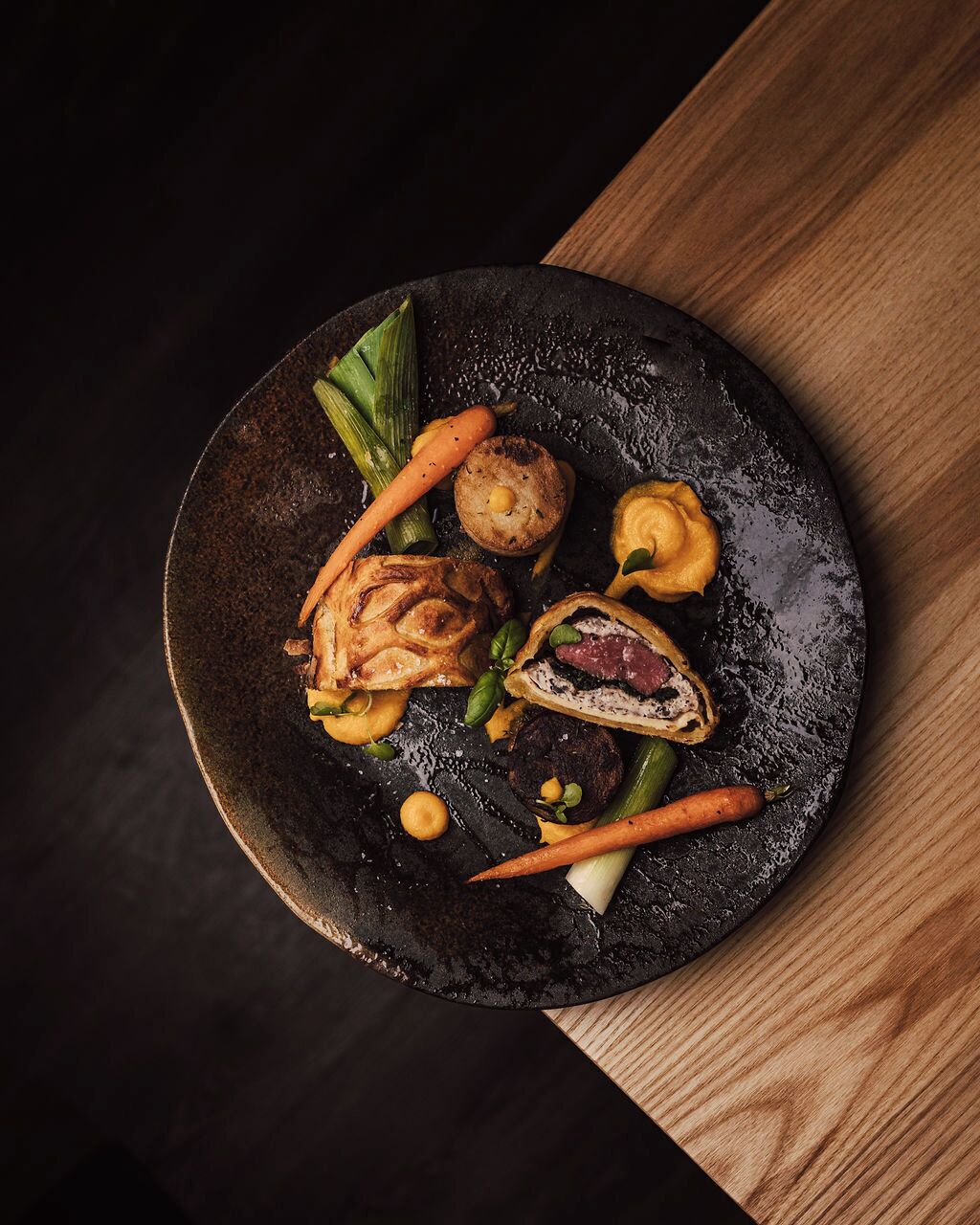 Ein schwarzer Teller mit verschiedenen Speisen, darunter Fleisch, Gemüse und ein Gebäckstück.